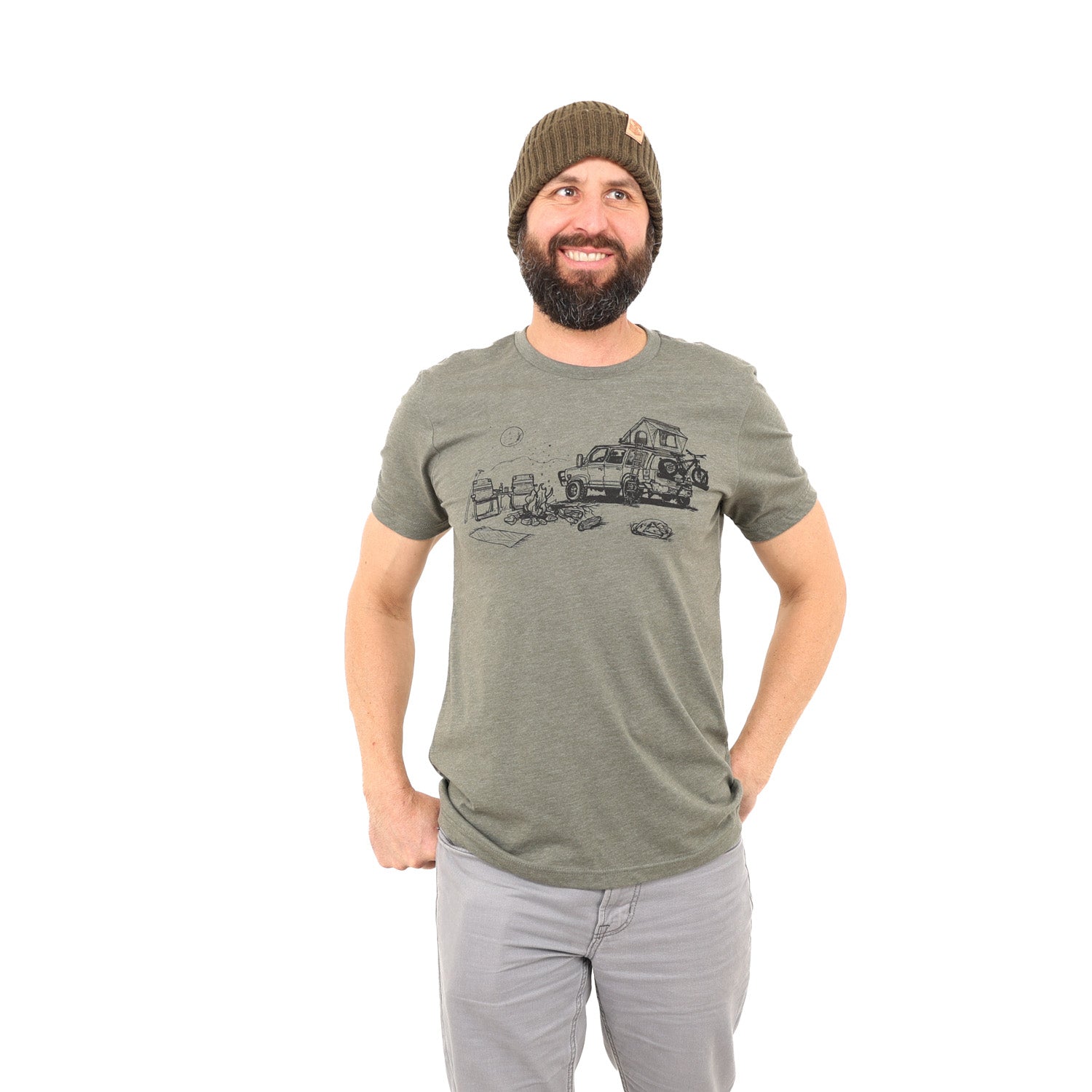 Man wearing army green shirt with black ink print of campfire scene; car with rooftop tent with ladder, bike on the back. Campfire, two chairs, mountains and full moon.