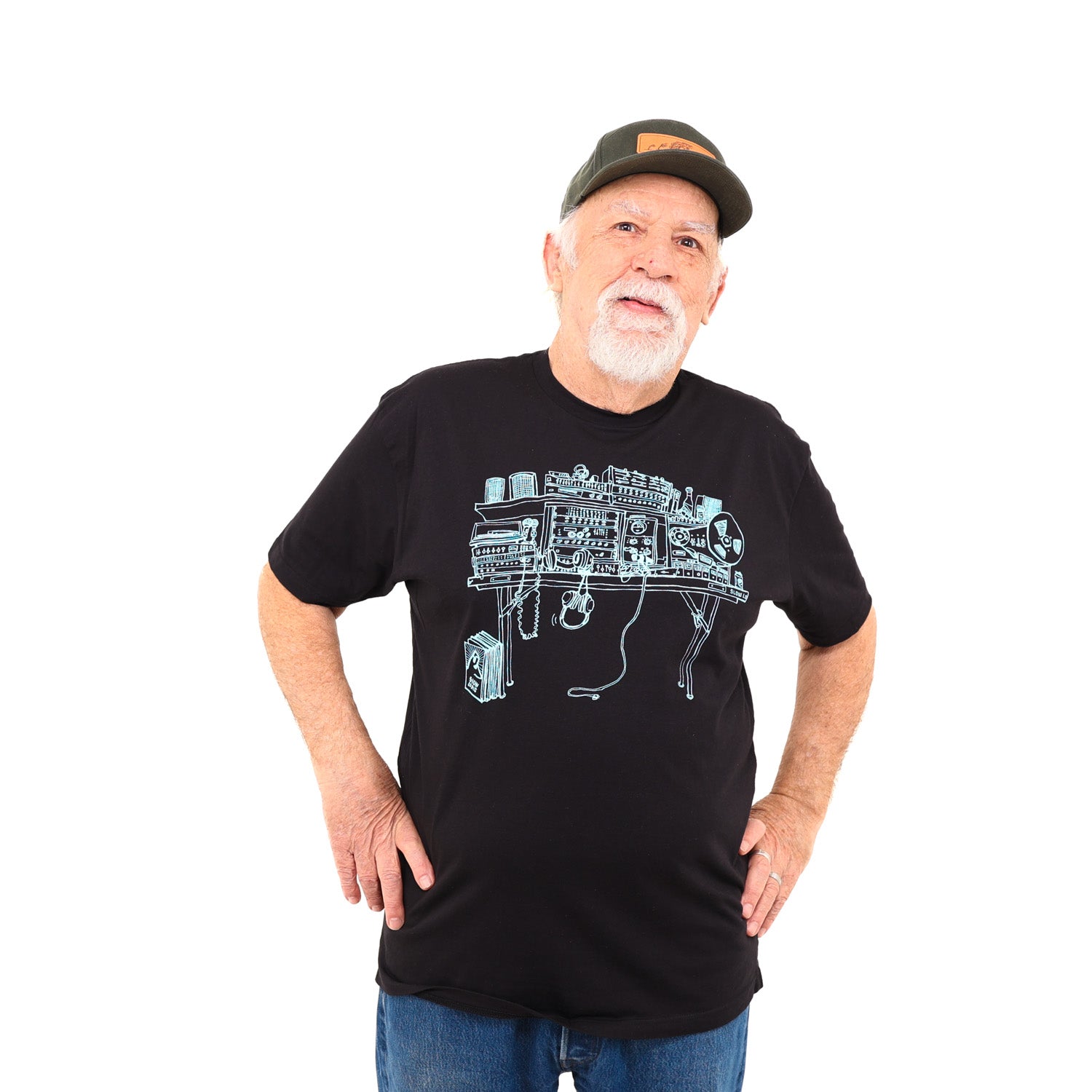 Man wearing hat standing with hands on his hips in a white background. Man is wearing black t-shirt with light blue ink of a recording studio