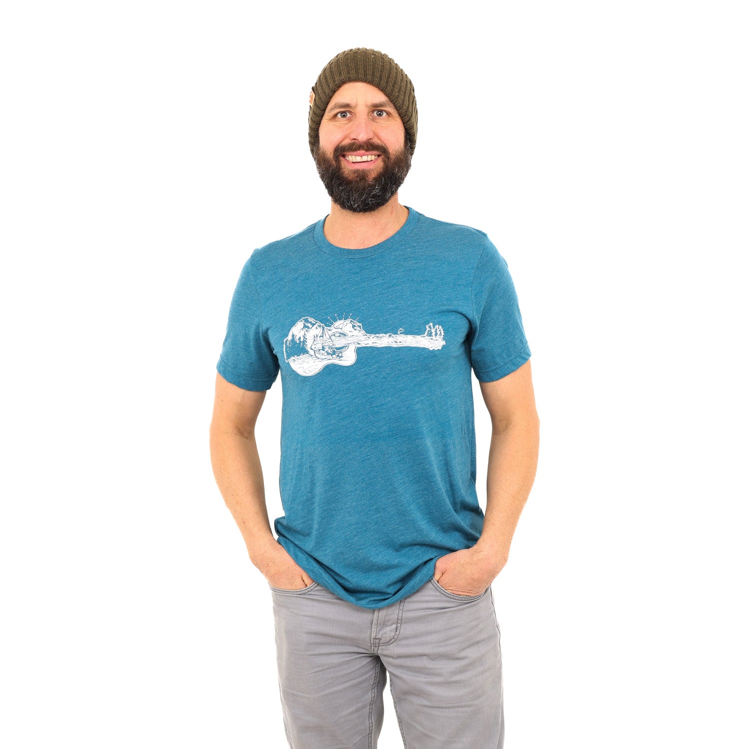 Man standing in white background with his hands in his pockets. Wearing grey jeans and a blue t-shirt with a guitar on it that actually a mountain boat scene.