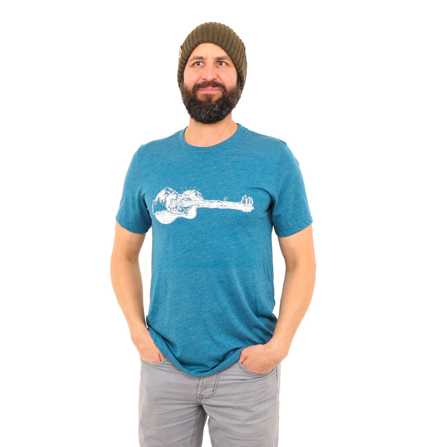 Man standing in white background with his hands in his pockets. Wearing grey jeans and a blue t-shirt with a guitar on it that actually a mountain boat scene.