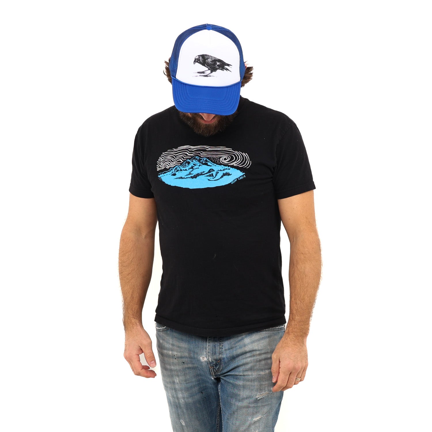 Man looking down wearing a ball cap in blue jeans and a black t-shirt with a blue mountain and white wind lines screenprinted