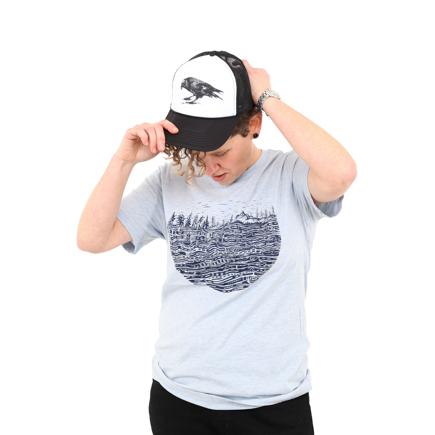 Woman putting a ball cap on while wearing heather light shirt with black ink print of the salish sea with mountains, trees, sky in the background.