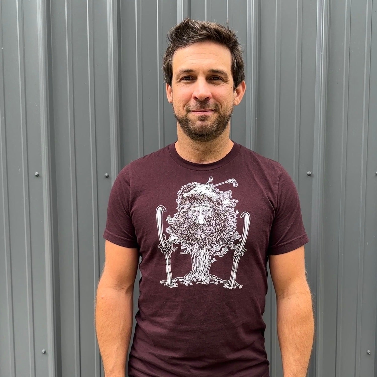Man wearing maroon t shirt with white print of close up of tree man holding skis with ski poles in his top branches