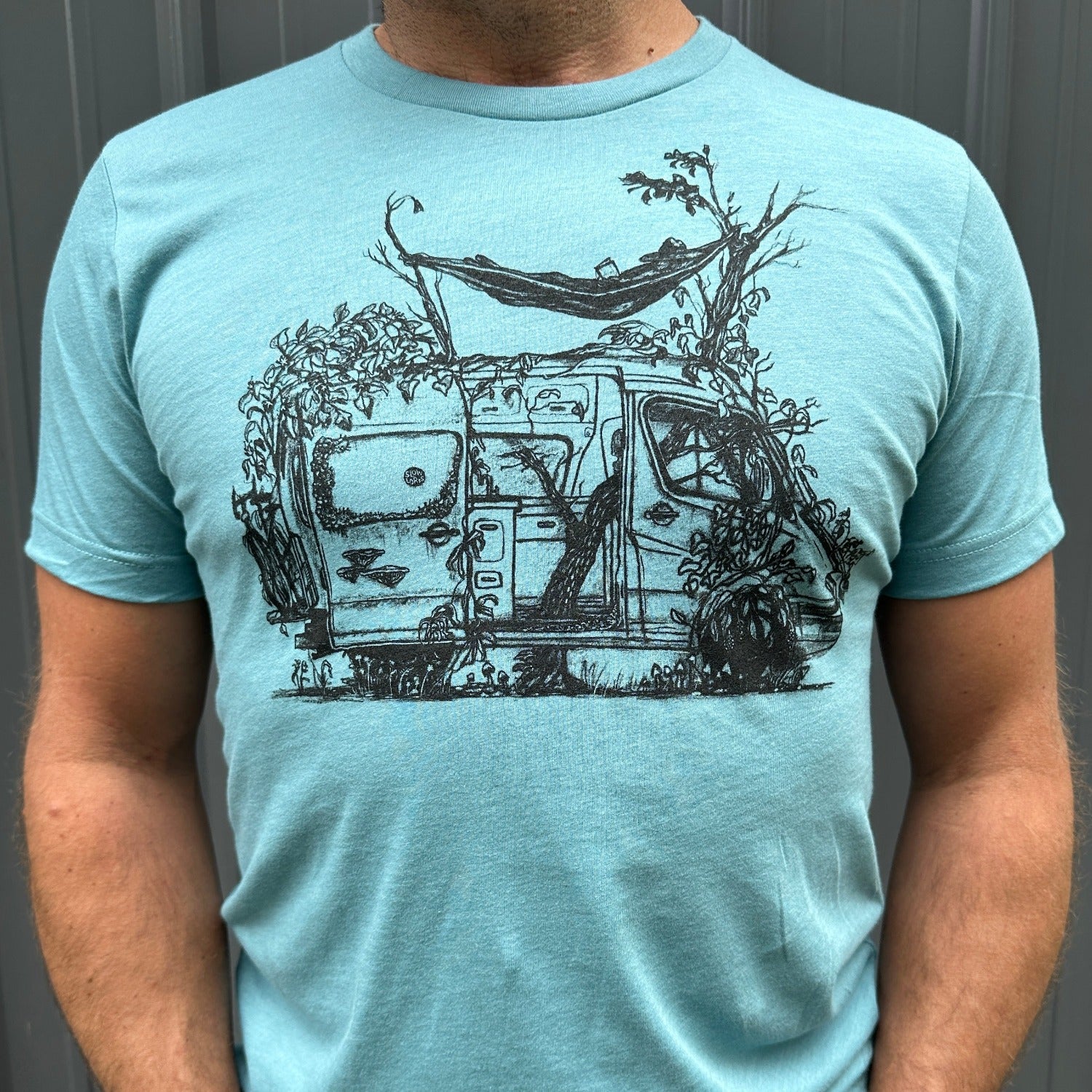 blue t-shirt with delapadated camper being taken back by earth. Tree growing right up the center of the van with person laying in hammock above sprinter like van