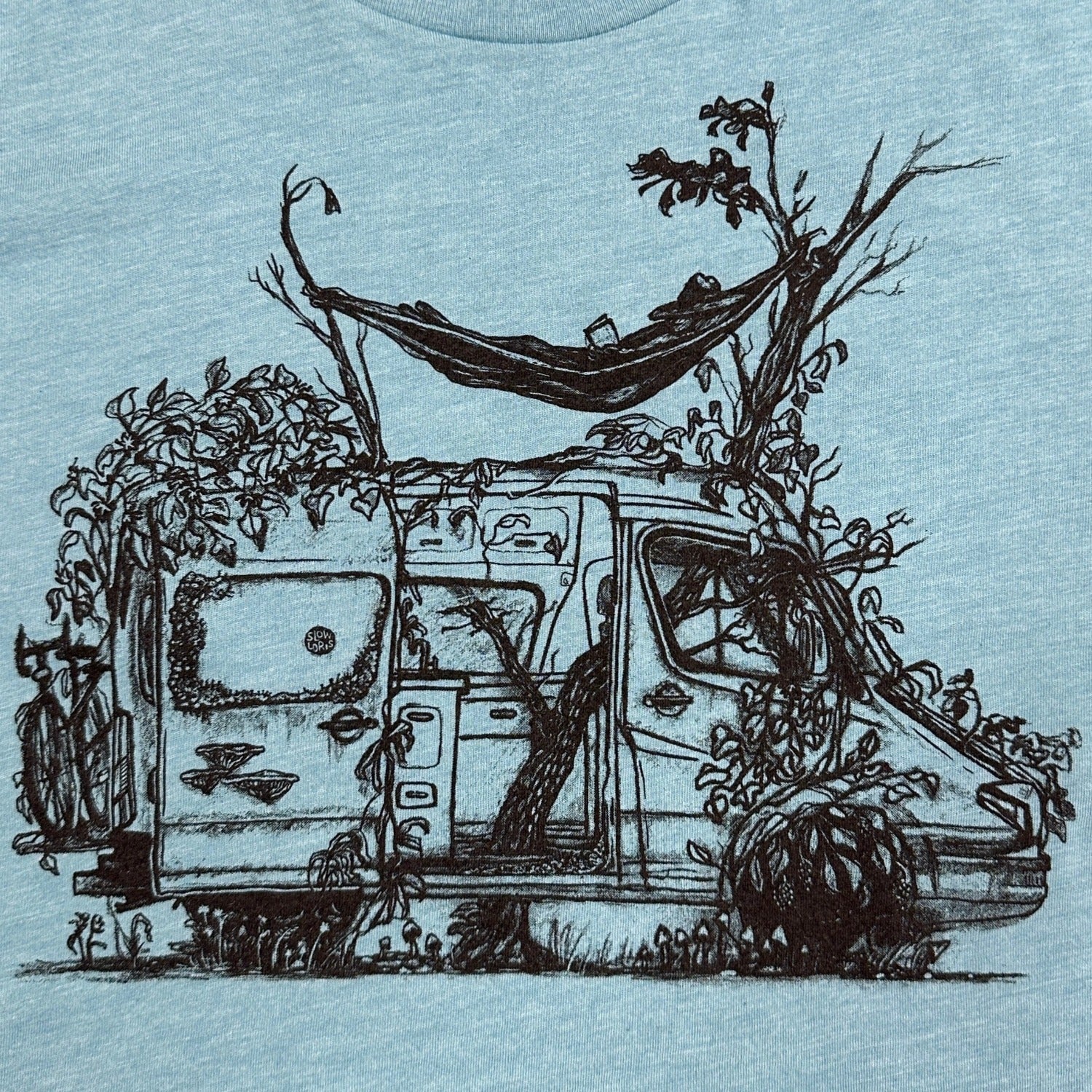 blue t-shirt with delapadated camper being taken back by earth. Tree growing right up the center of the van with person laying in hammock above sprinter like van