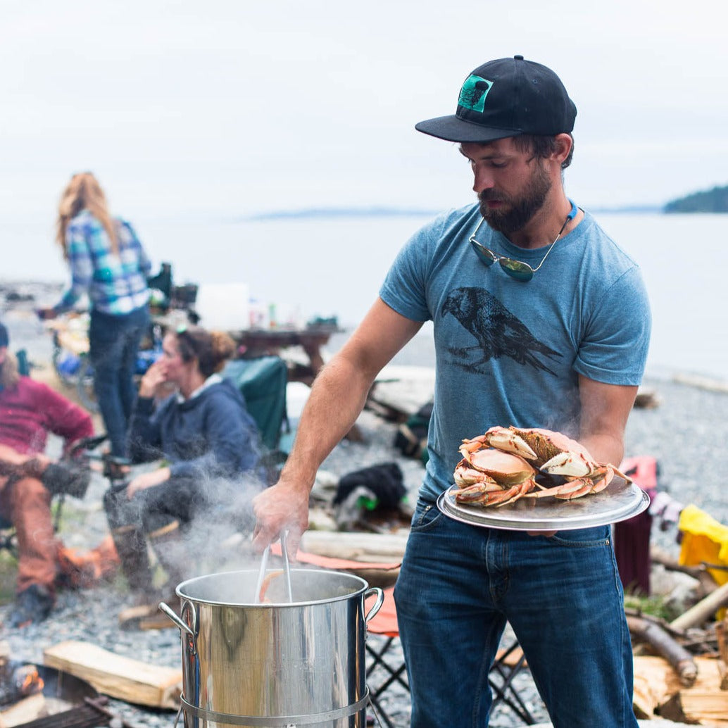 man pulling cooked crab out of a pot while wearing a blue shirt with a screen printed crow on it.