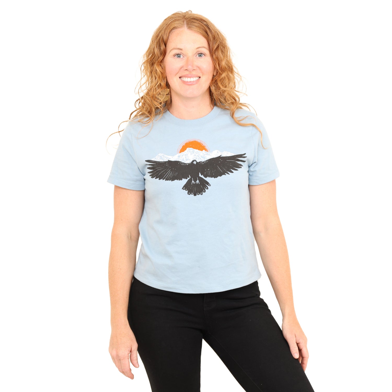 woman wearing light blue shirt with a raven wings spread and mountains and son above.