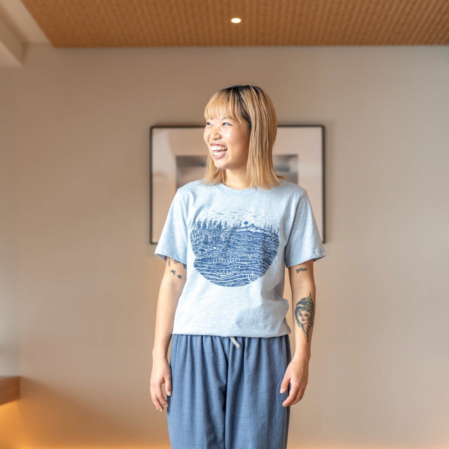 Smiling woman wearing heather light shirt with black ink print of the salish sea with mountains, trees, sky in the background.