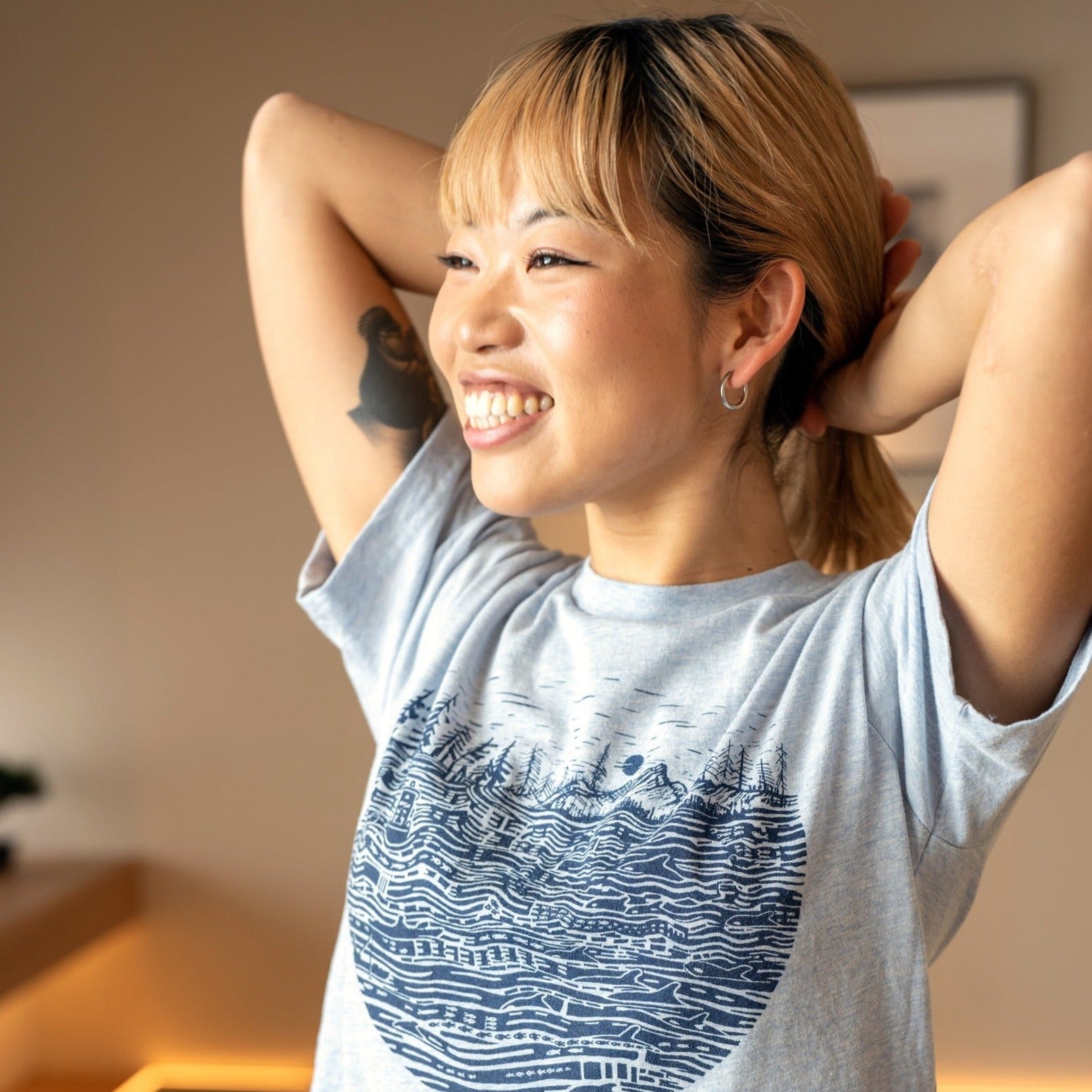 Smiling woman wearing heather light shirt with black ink print of the salish sea with mountains, trees, sky in the background.