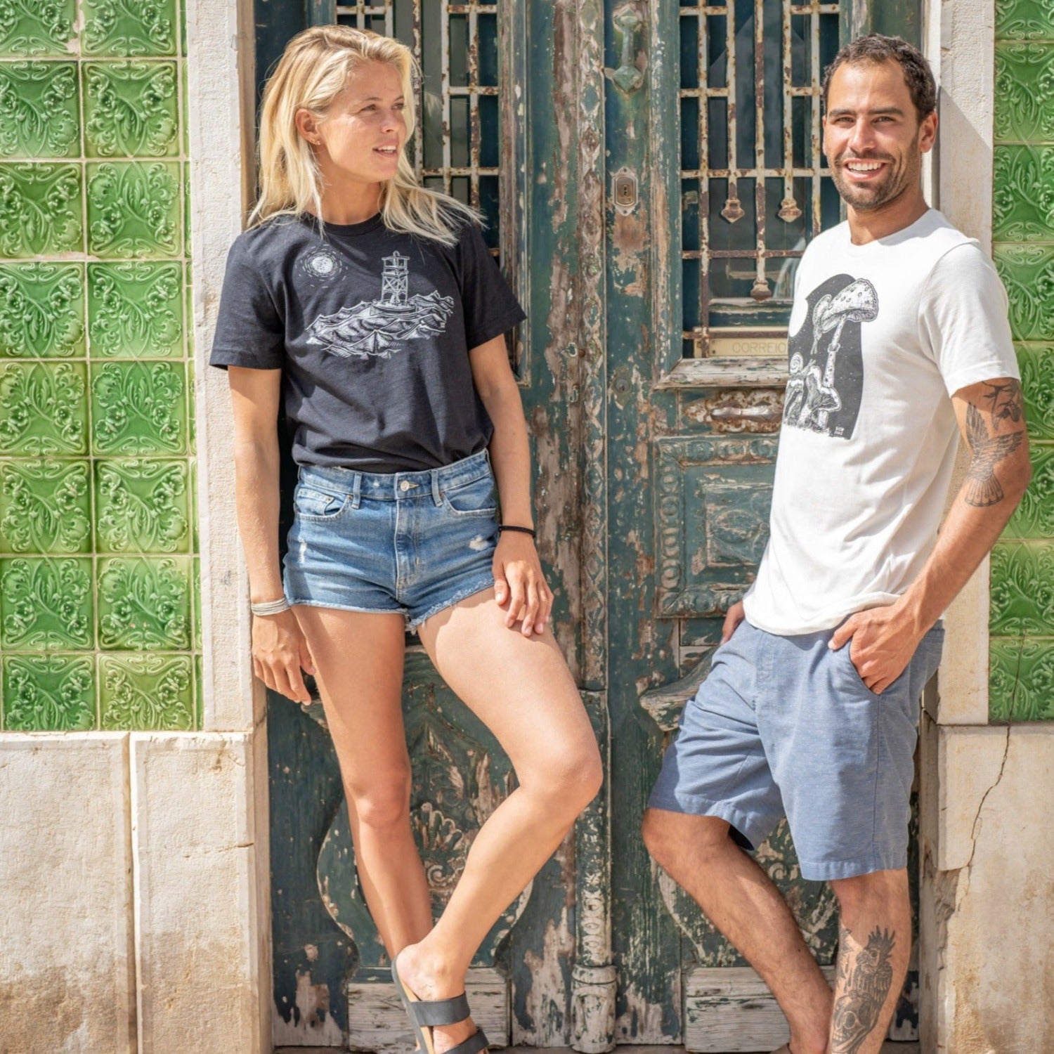 man and woman couple standing in front of beautiful old door together. Woman is wearing a black t-shirt with white ink of stormy seas and a rocking channel marker and a moon/sun in the distance. Man is wearing a Slow Loris Full Moon Buffet t-shirt.