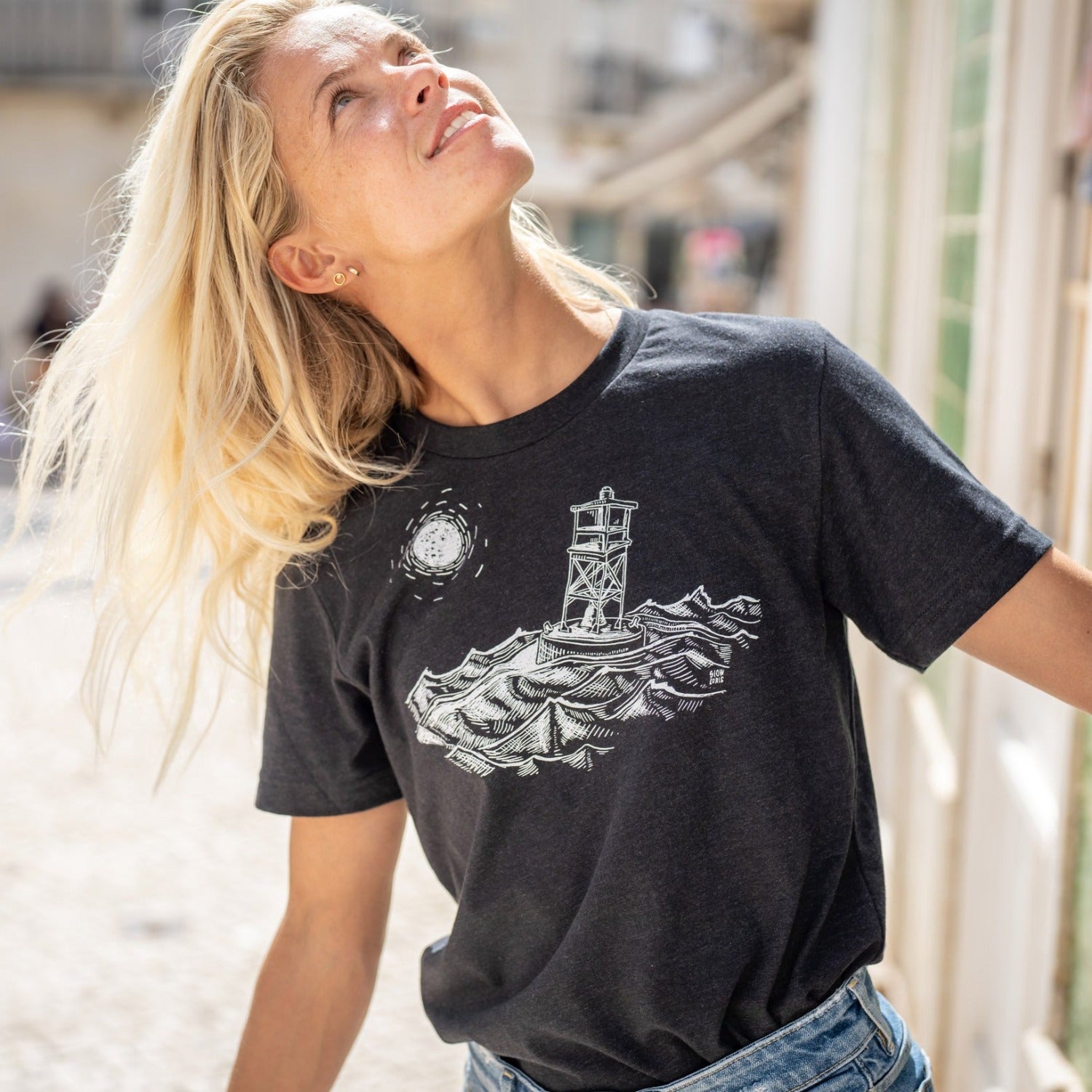 woman looking up while wearing black t-shirt with white ink of stormy seas and a rocking channel marker and a moon/sun in the distance.