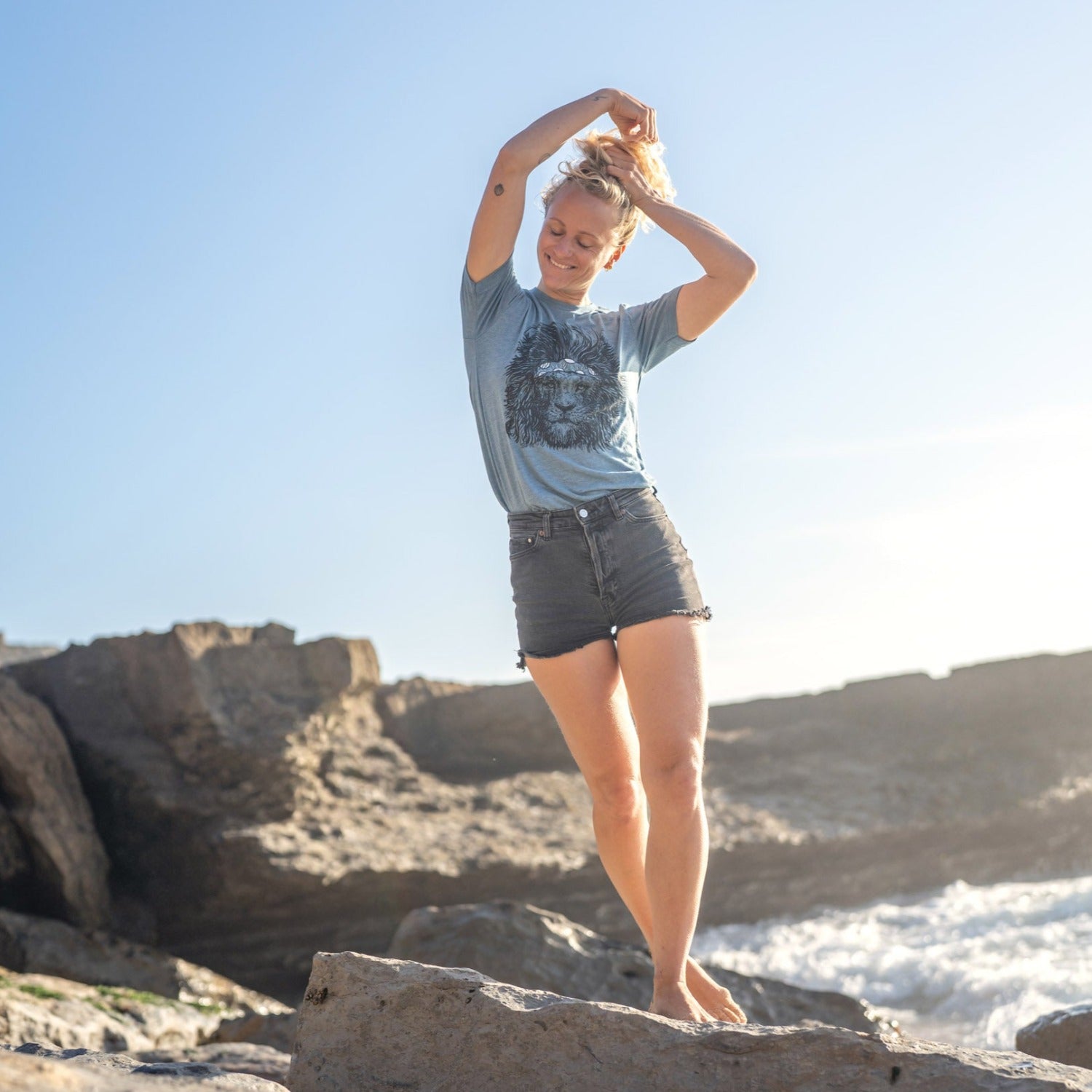Woman on beach wearing a steal blue tee with a screen printed lion wearing a headband on it.