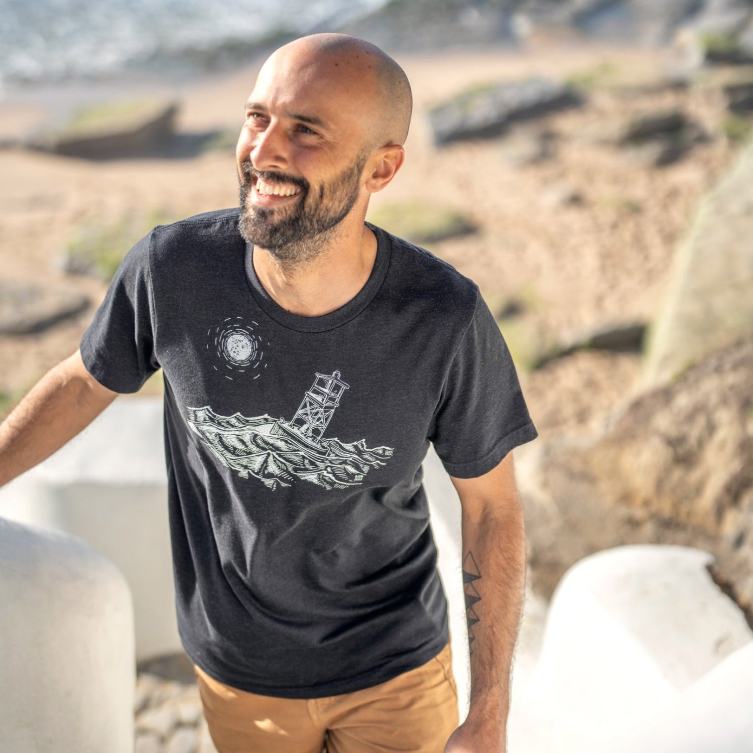 Man walking up stairs from the beach wearing black t-shirt with white ink of stormy seas and a rocking channel marker and a moon/sun in the distance.