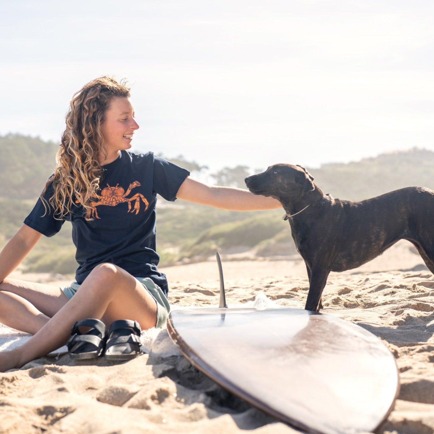 Girl sitting on beach with surfboard while petting dog. She is wearing a blue tee with a Dungeness crab printed on it.