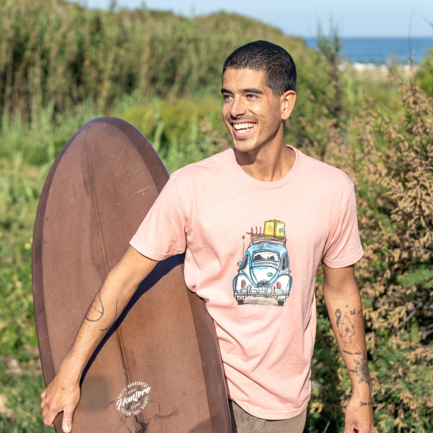 Man carrying surfboard while wearing pink shirt with a VW bug on it. 