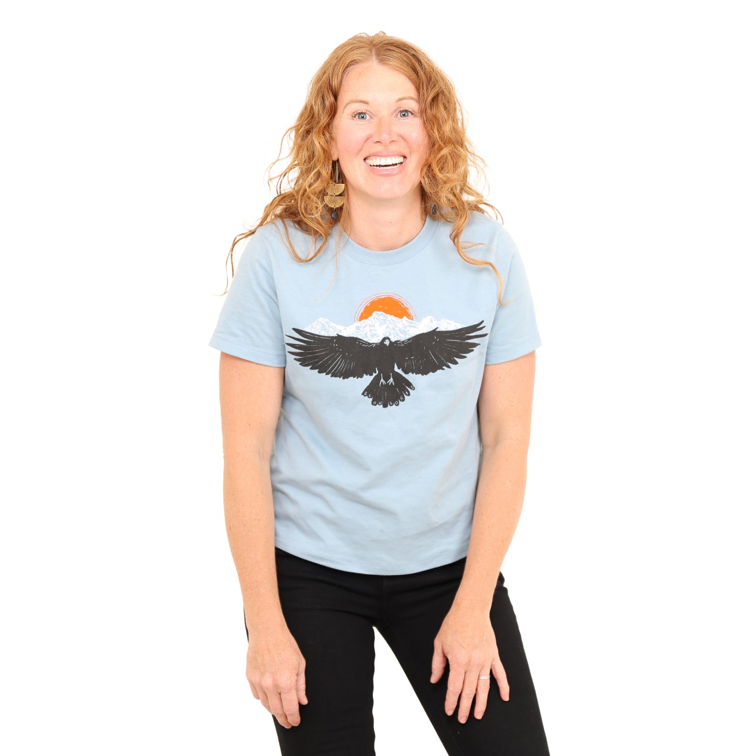 woman wearing light blue shirt with a raven wings spread and mountains and son above.