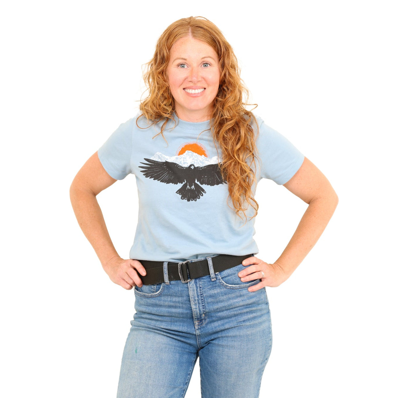 woman wearing light blue shirt with a raven wings spread and mountains and son above.