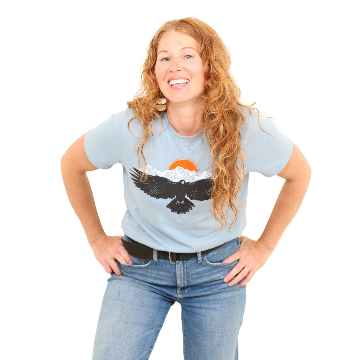 woman wearing light blue shirt with a raven wings spread and mountains and sun above.