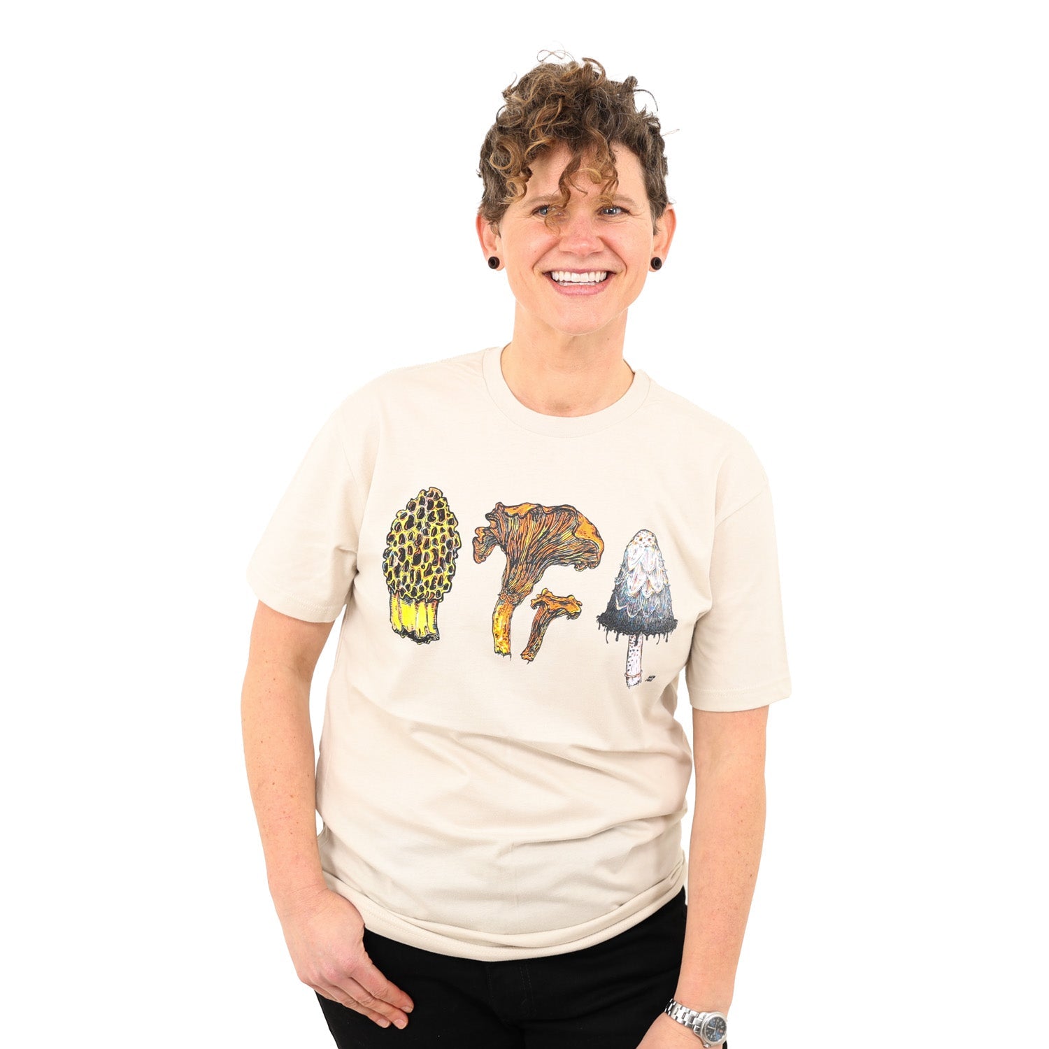 smiling woman wearing a tan colored shirt with three mushrooms- morel, chantrelle, and shaggy mane