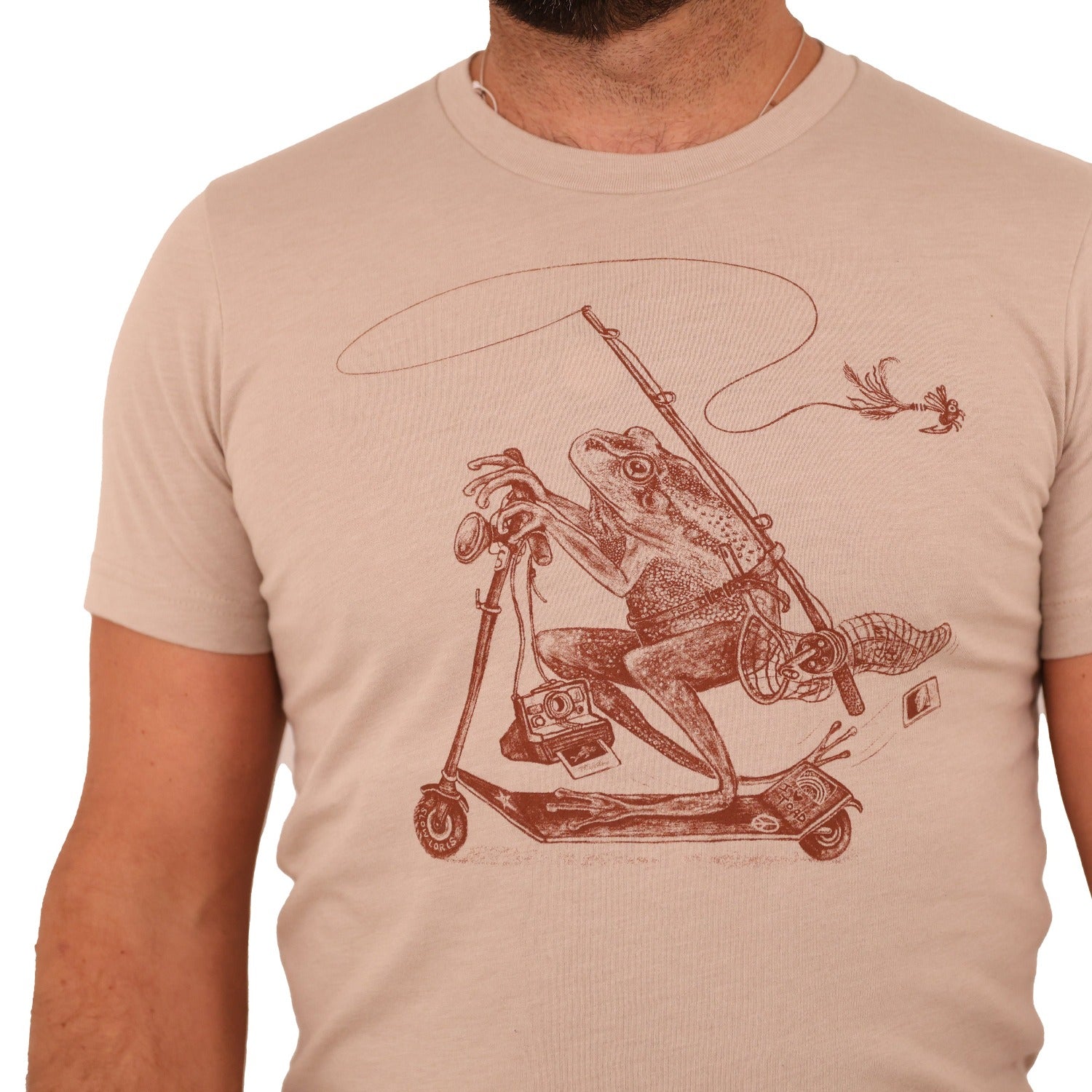 man wearing tan shirt with print of a frog riding a scooter with a fly fishing rod, net, polaroid camera