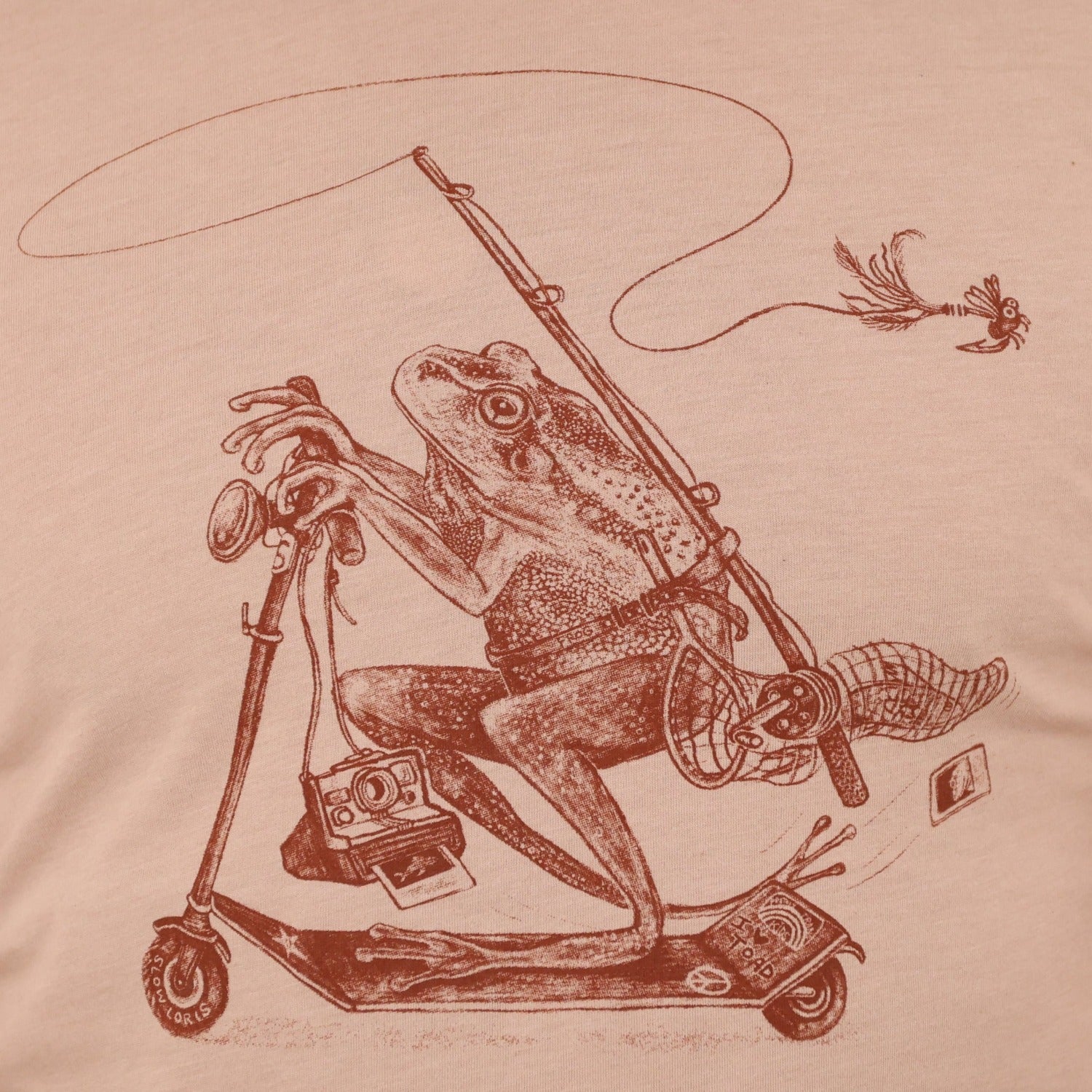 close up of shirt with print of a frog riding a scooter with a fly fishing rod, net, polaroid camera