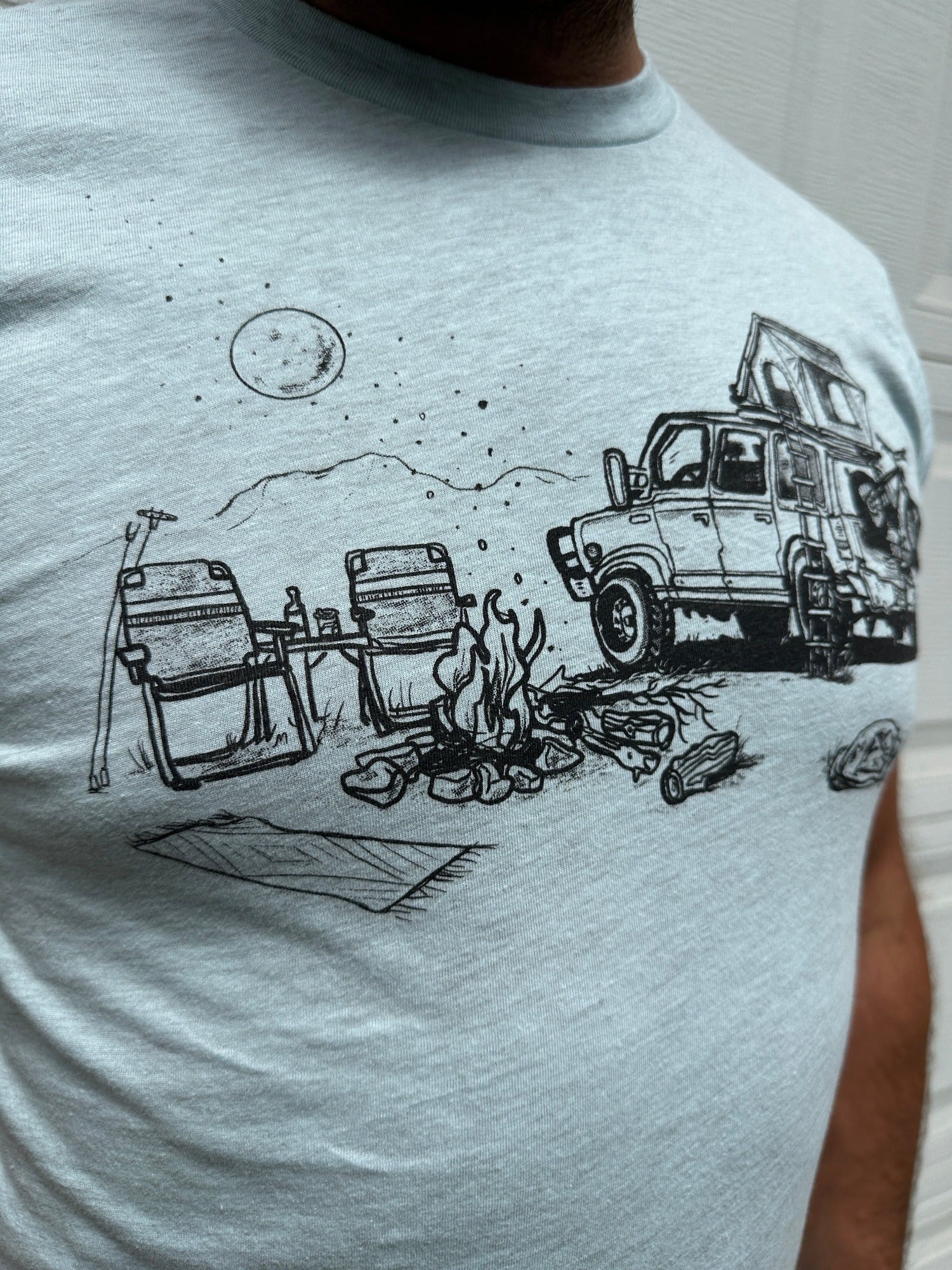 Man wearing light blue shirt with black ink print of campfire scene; car with rooftop tent with ladder, bike on the back. Campfire, two chairs, mountains and full moon.