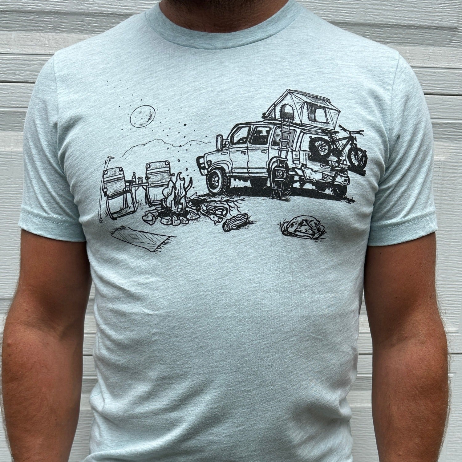 Man wearing light blue shirt with black ink print of campfire scene; car with rooftop tent with ladder, bike on the back. Campfire, two chairs, mountains and full moon.