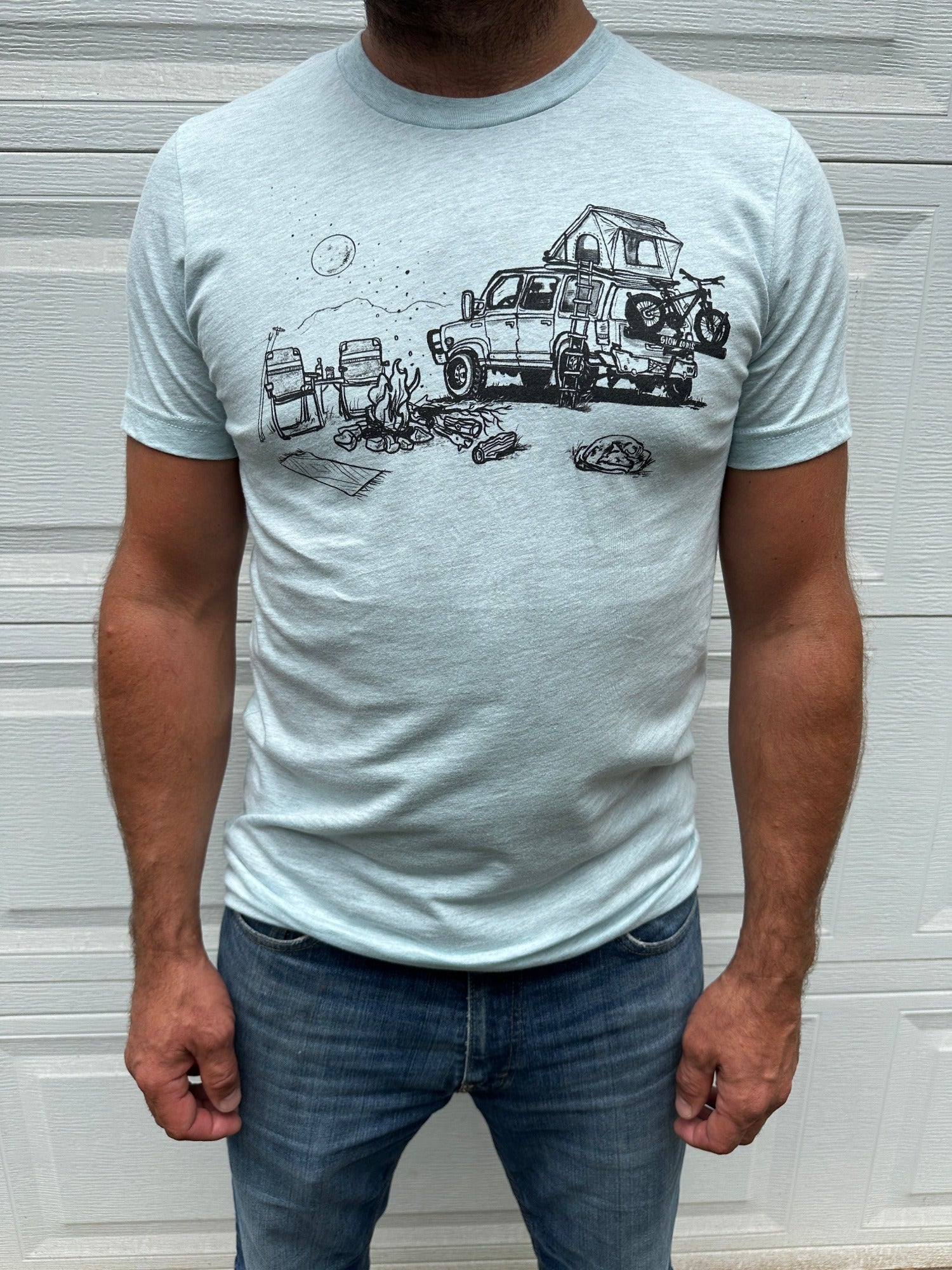 Man wearing light blue shirt with black ink print of campfire scene; car with rooftop tent with ladder, bike on the back. Campfire, two chairs, mountains and full moon.