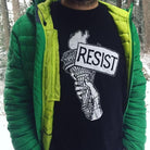 man outside with snowy background wearing black resist t shirt and jeans with a green puffy zip up 