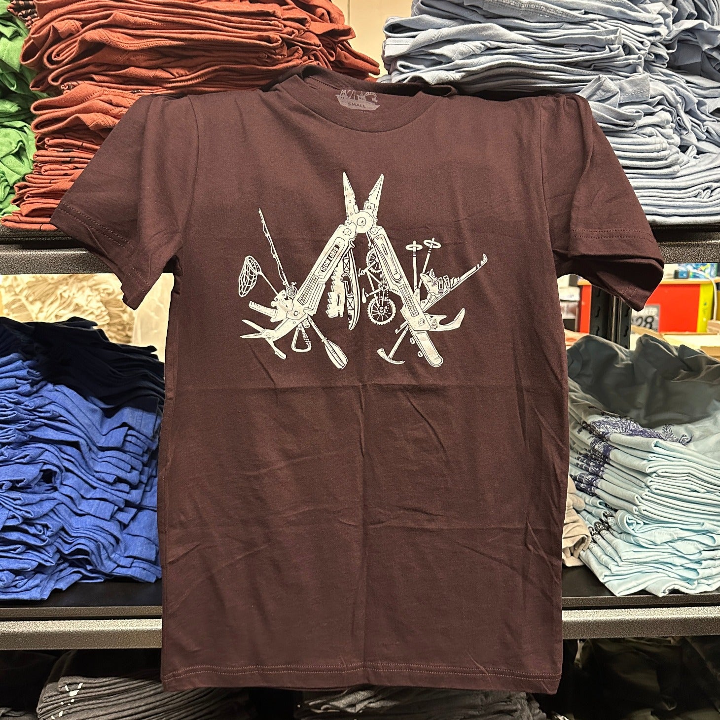 oxblood colored t-shirt with a white print of a multitool. The tools within the multitool are actually adventuring items- kayak, fishing pole, skis, ice axe, etc etc.