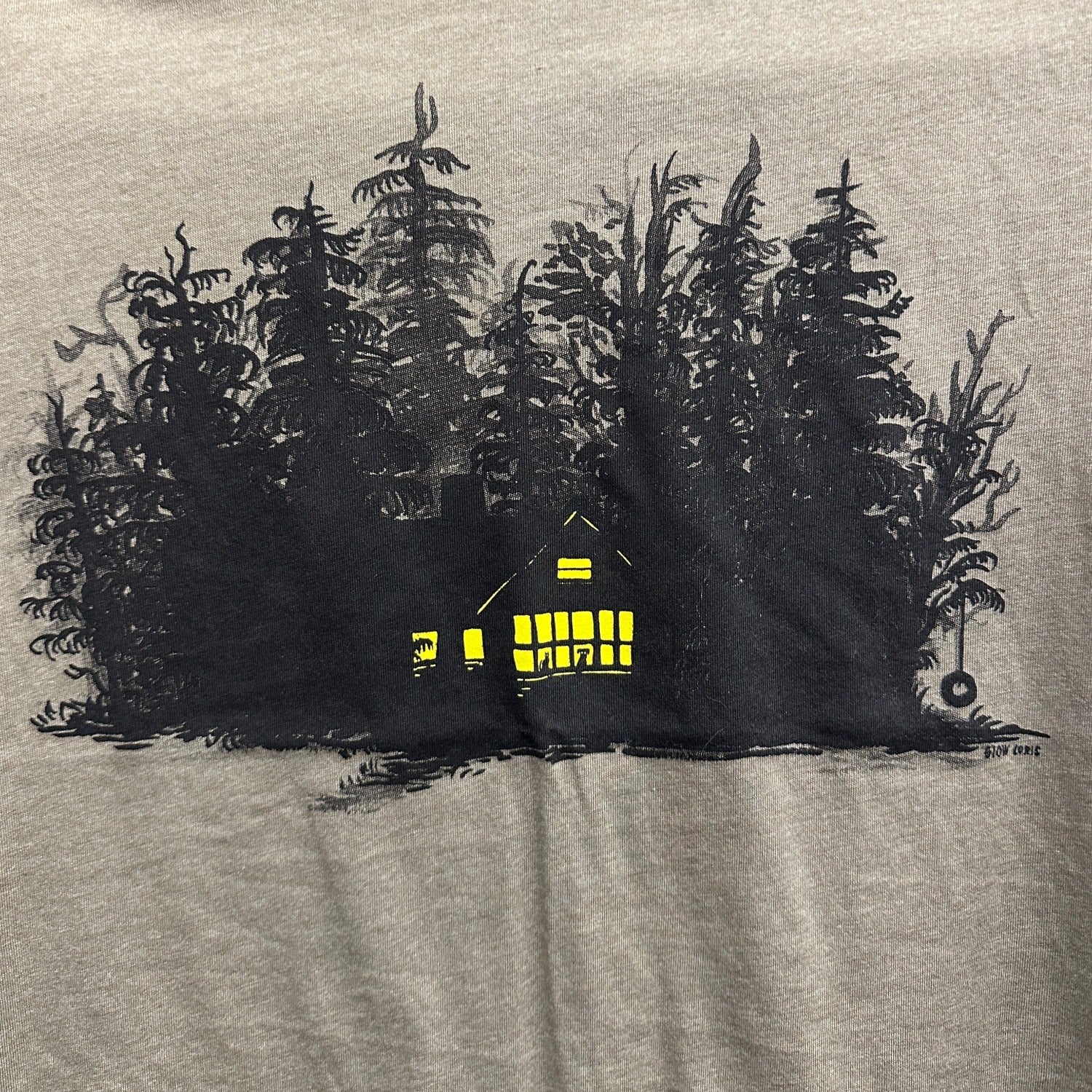 close up showing print of brown t-shirt with print of black ink of silhouetted trees with yellow ink house lights shining out of darkness. Two animals sitting in the window.