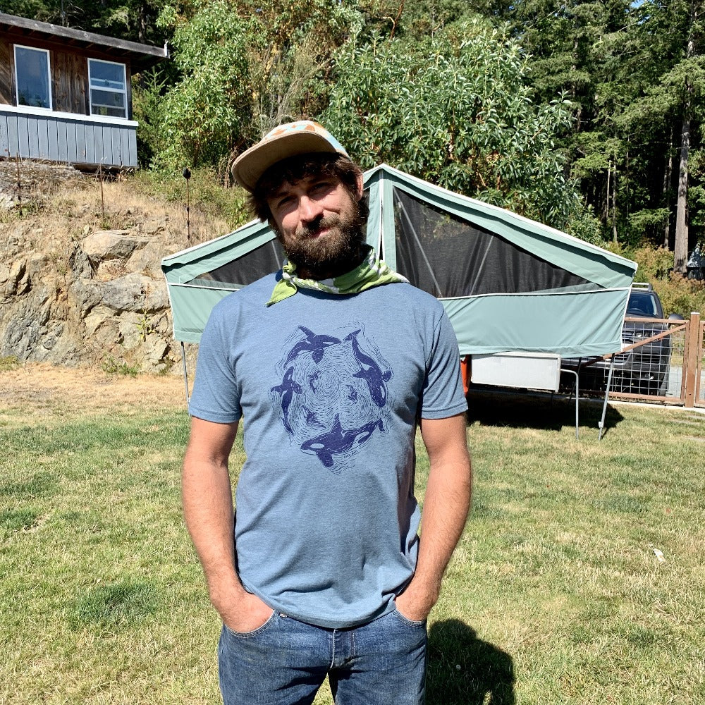 Bearded man wearing a hat and a hankie with light blue shirt and dark blue print of orcas swimming in a circle