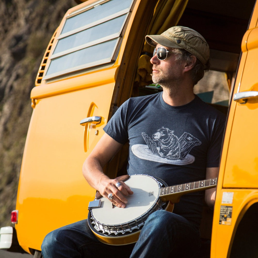 Man sitting in a VW van slider door playing a banjo looking off into the distance while wearing a blue t shirt with white screen printed in of a bear sitting on a floatie in the water.