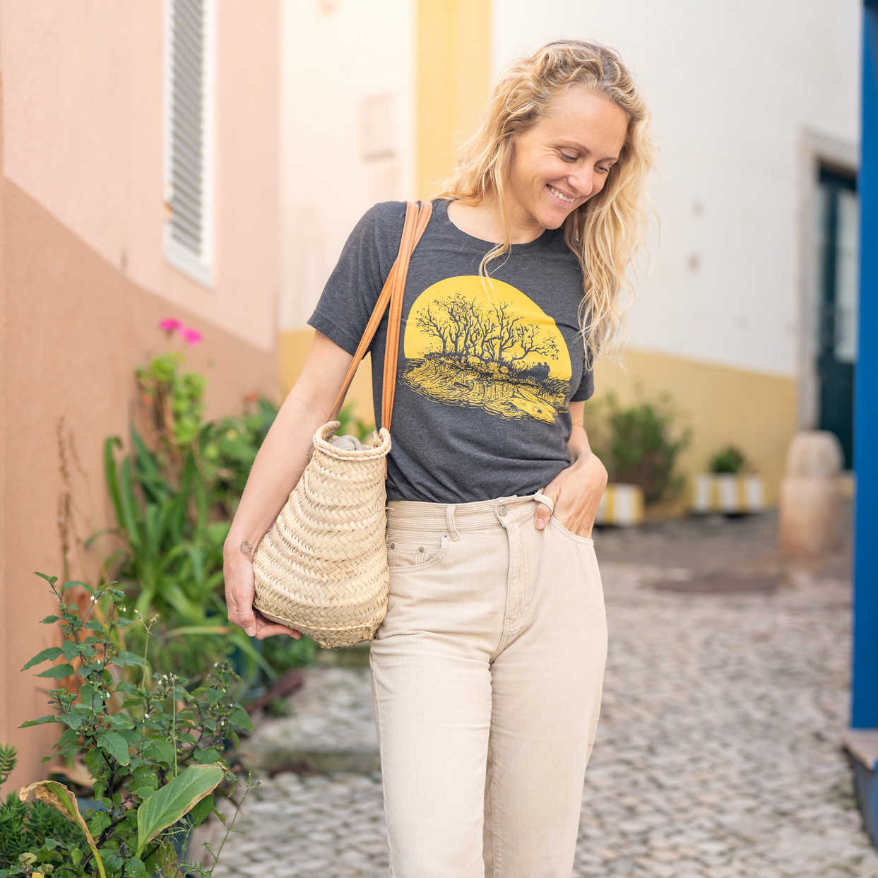 Woman walking with bag on her shoulder and one hand in her pocket. Girl is wearing light tan jeans and a dark grey t-shirt with yellow print of kayakers on the beach of a tiny island watching the sunset