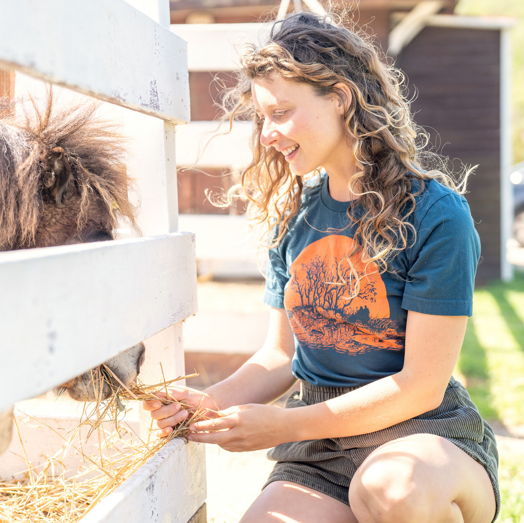 Woman feeding donkey while wearing a blue t-shirt with orange print of a tiny island with two kayaks on the beach