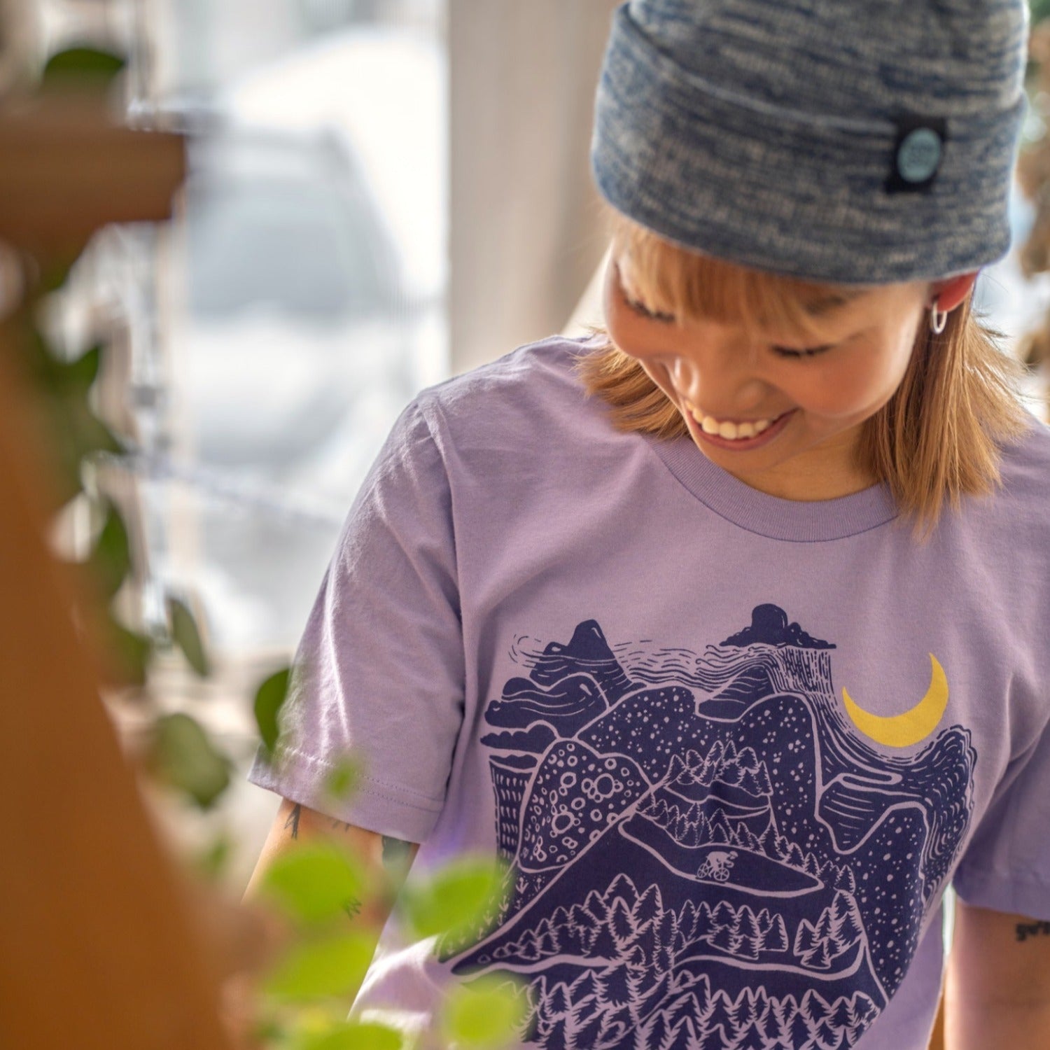 Woman wearing light purple shirt with dark blue ink. Mountain scene with raincloud, crescent moon, and bicyclist riding down the mountain