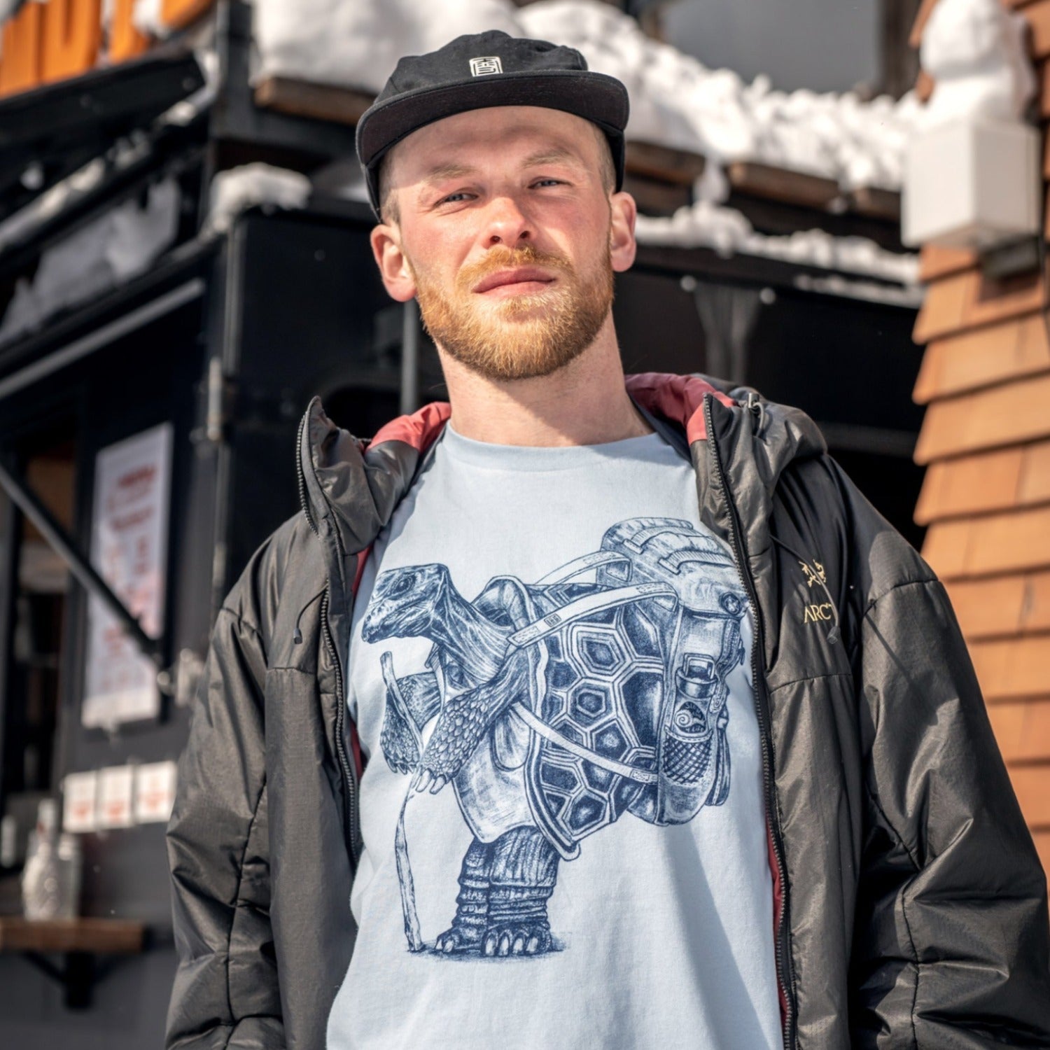 Man standing outside with snow on the buildings in the background. Man is wearing a ball cap, jacket, light blue shirt with dark blue ink of a hiking tortoise.