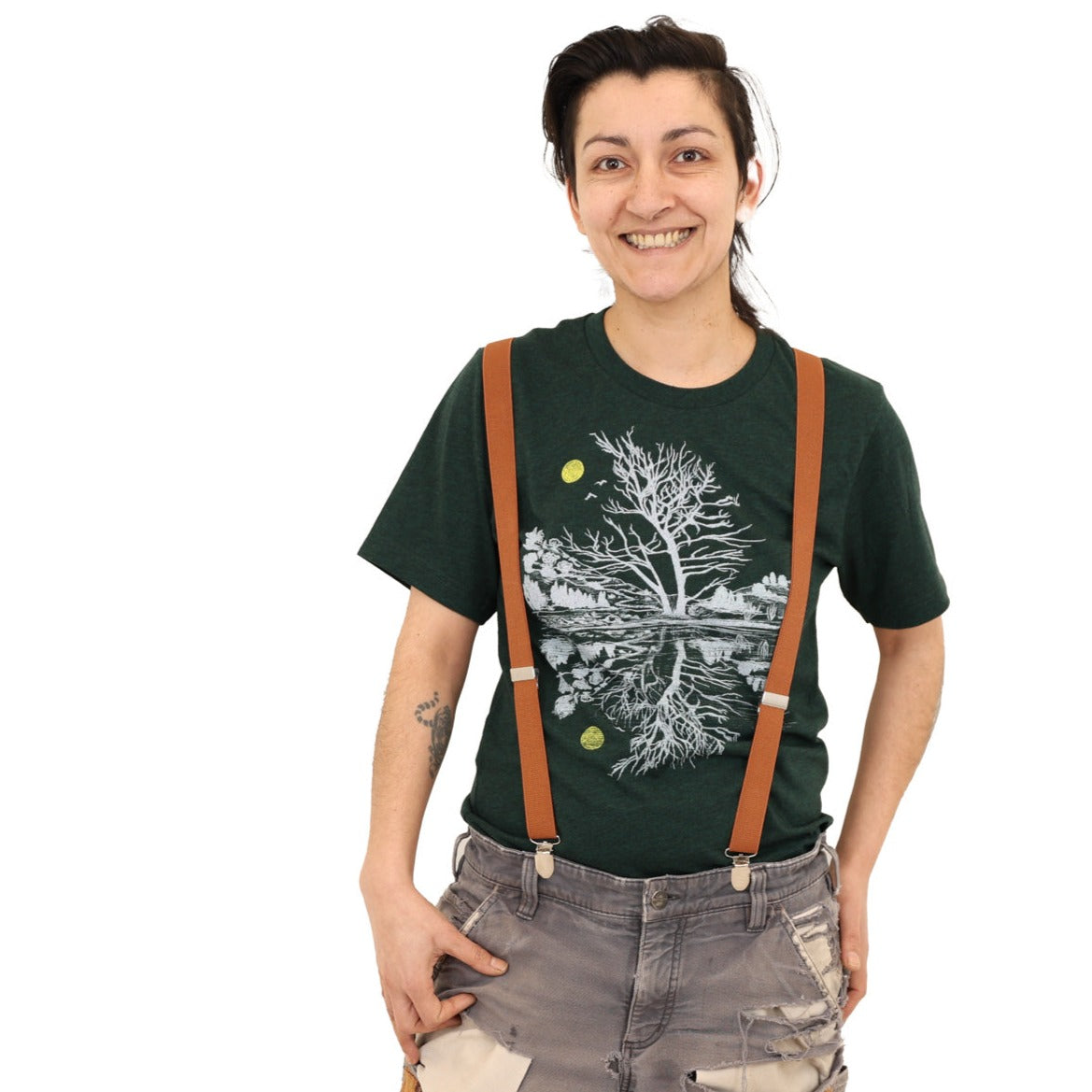 woman wearing suspenders and patchy pants with a green t-shirt with light ink and a yellow moon with scene reflecting over water.