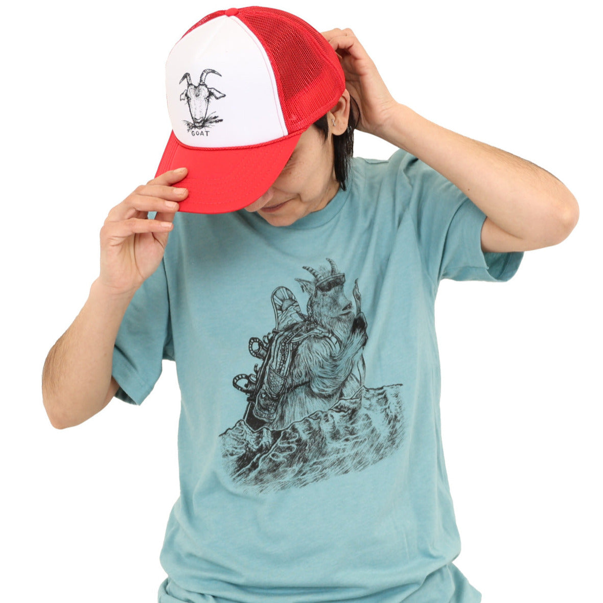 woman wearing soft light t-shirt with black ink of a mountain goat hiking with a walking stick, sunglasses, and a snowboard on his backpack. Mountains with fresh tracks anterior