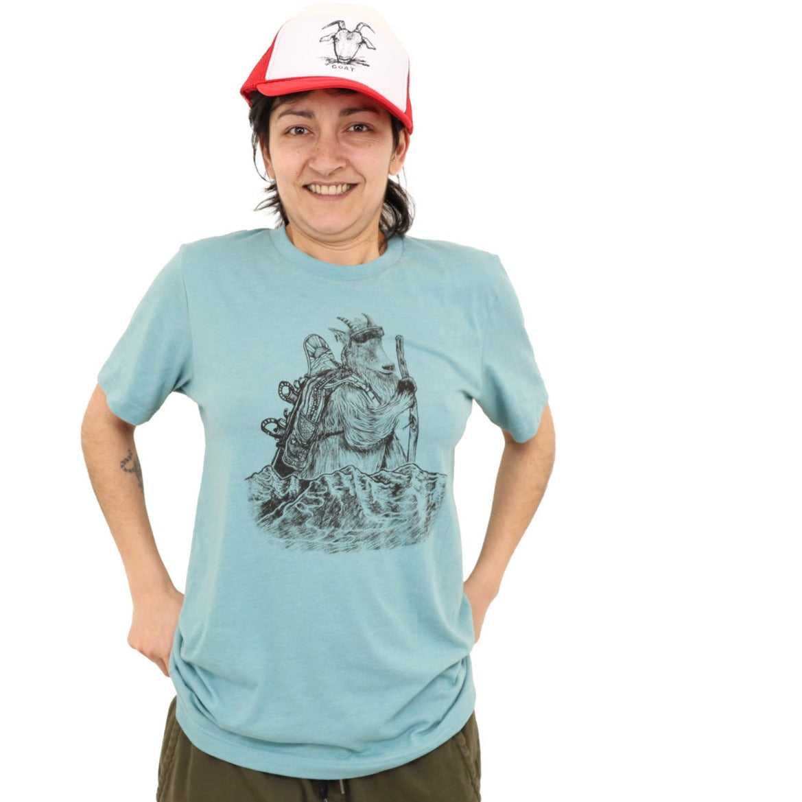 Woman wearing soft light t-shirt with black ink of a mountain goat hiking with a walking stick, sunglasses, and a snowboard on his backpack. Mountains with fresh tracks anterior