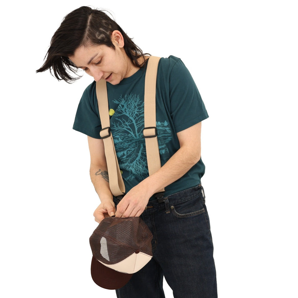 Woman wearing atlantic blue t-shirt with suspenders and a hat in her hands. Print is in a light blue tree & reflection over water with a yellow moon.