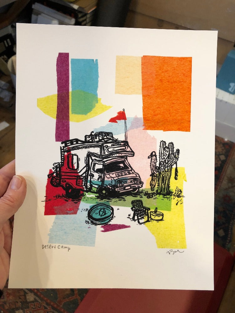 A hand holding our Desert Camp art print, a colorful tissue and ink painting of a truck camper parked next to a cactus and lawn chair.