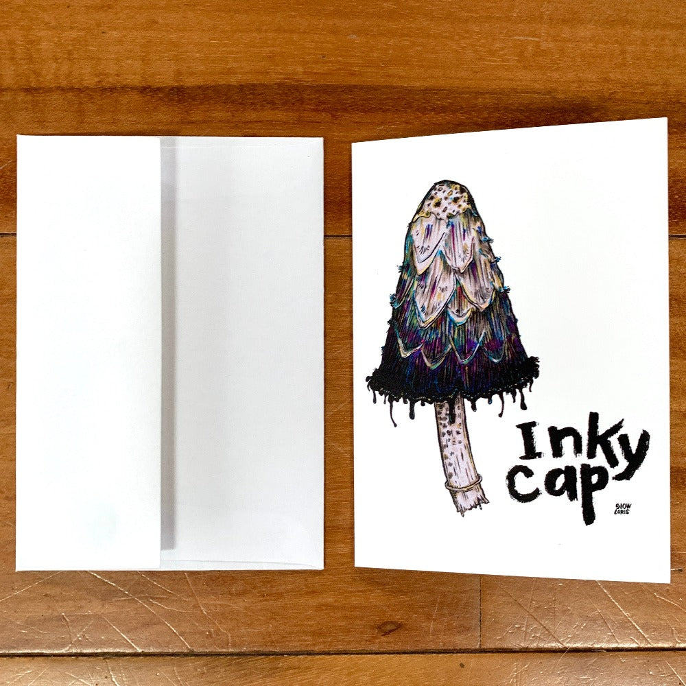A greeting card printed with a painting of an inky cap mushroom and the words Inky Cap in hand-lettering. The card is lying flat on a table next to an envelope.