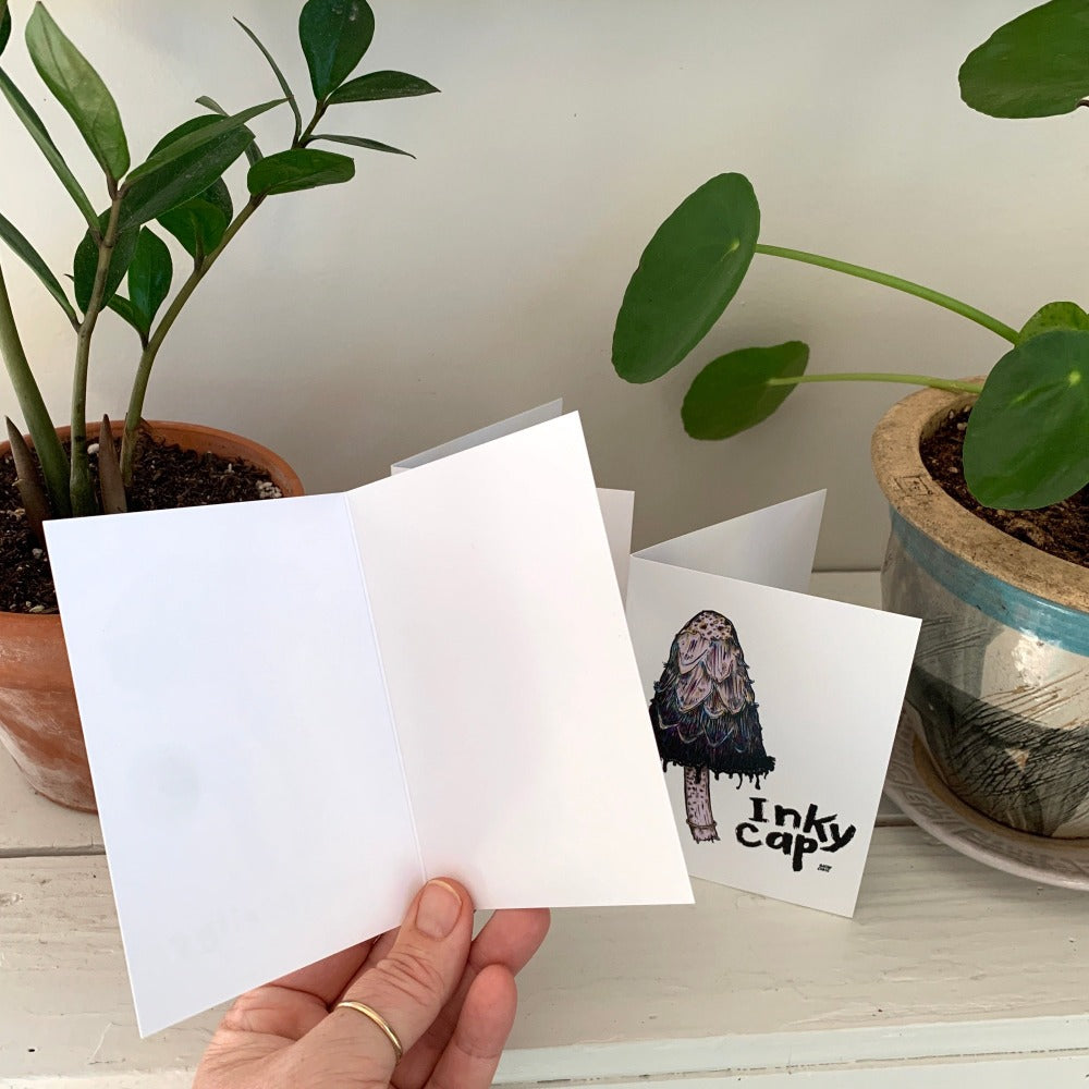 A hand holding a greeting card open to show that it is blank inside. In the background is a greeting card printed with an inky cap mushroom.