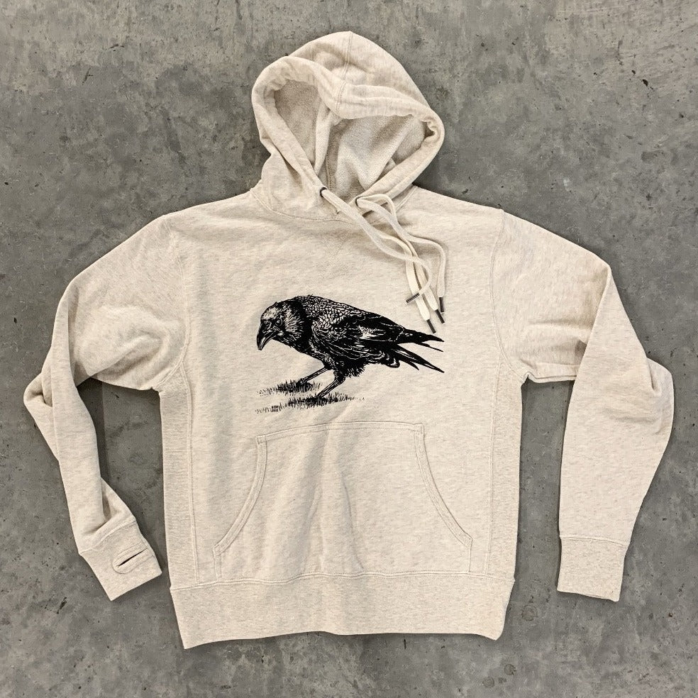 An oatmeal-colored hoodie, screen printed with a crow in black ink and laid flat against a gray background.