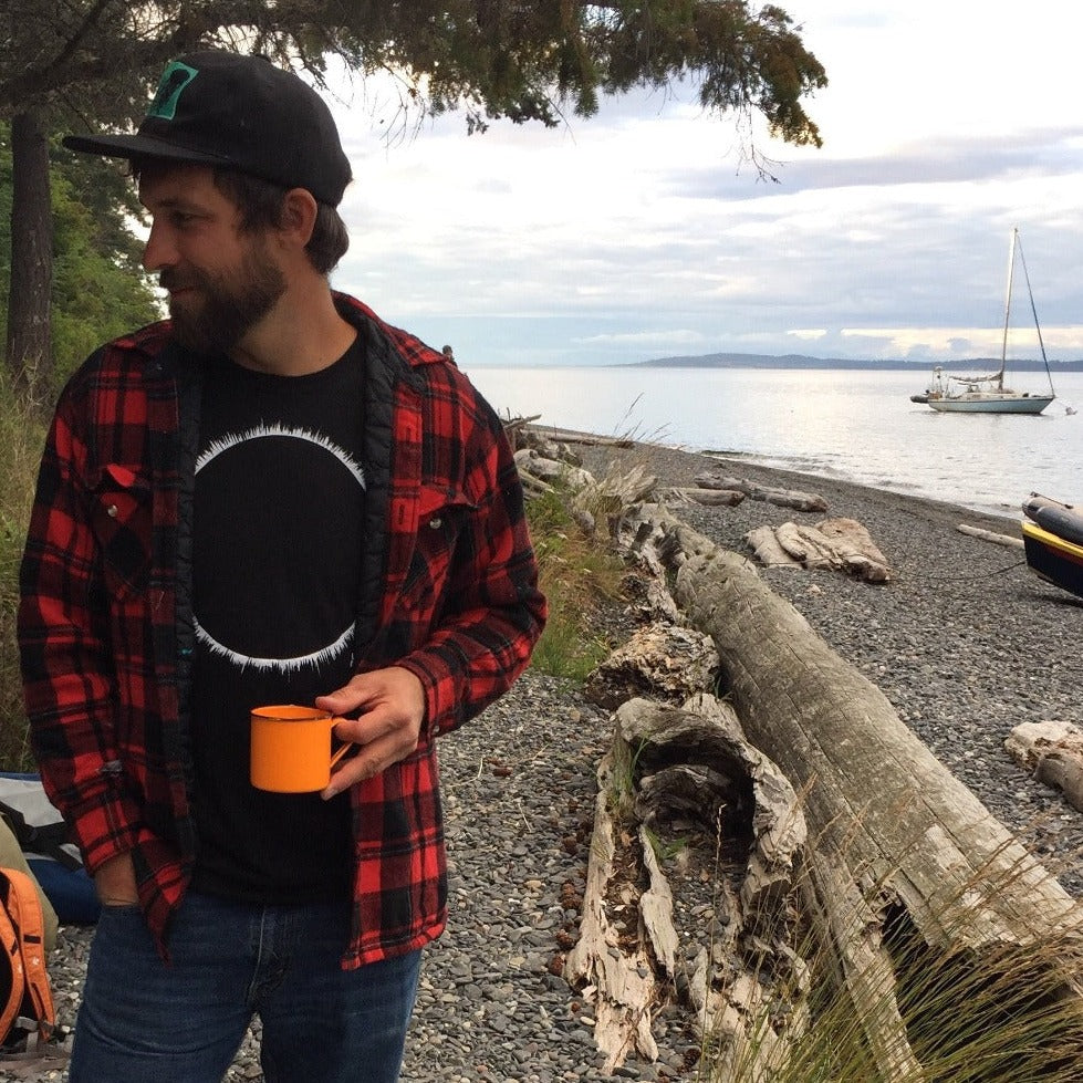 man on beach with sailboat in the background. Man is holding an orange cup talking with someone out of frame. Man is wearing a ball cap and a red flanel sweater that is open in the front showing the black t-shirt with white eclipse print on it.
