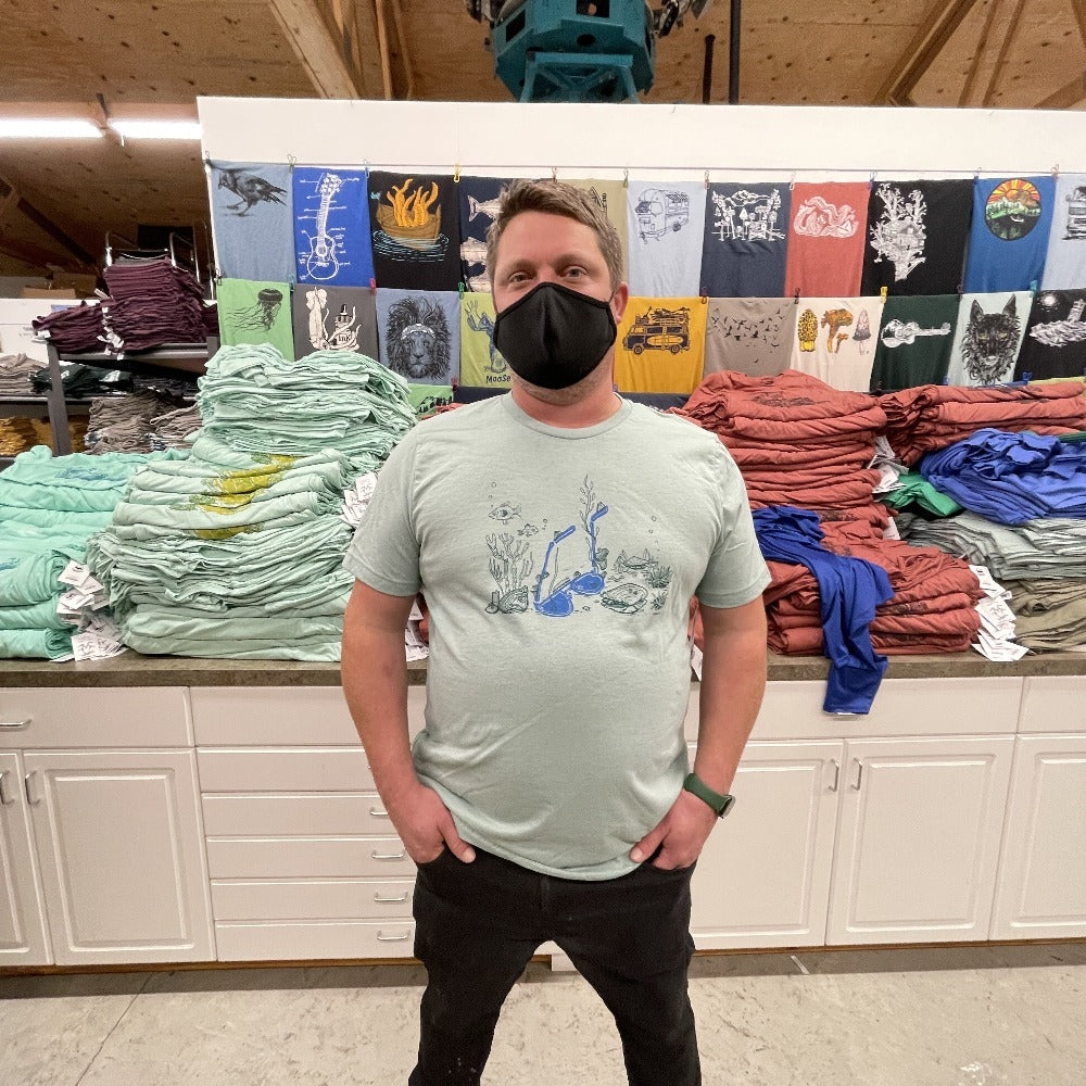 Man standing with hands in his pockets while wearing a mask on his face. Stacks of t-shirts behind. Man is wearing light soft blue t-shirt with print of sunglasses and sea creatures underwater
