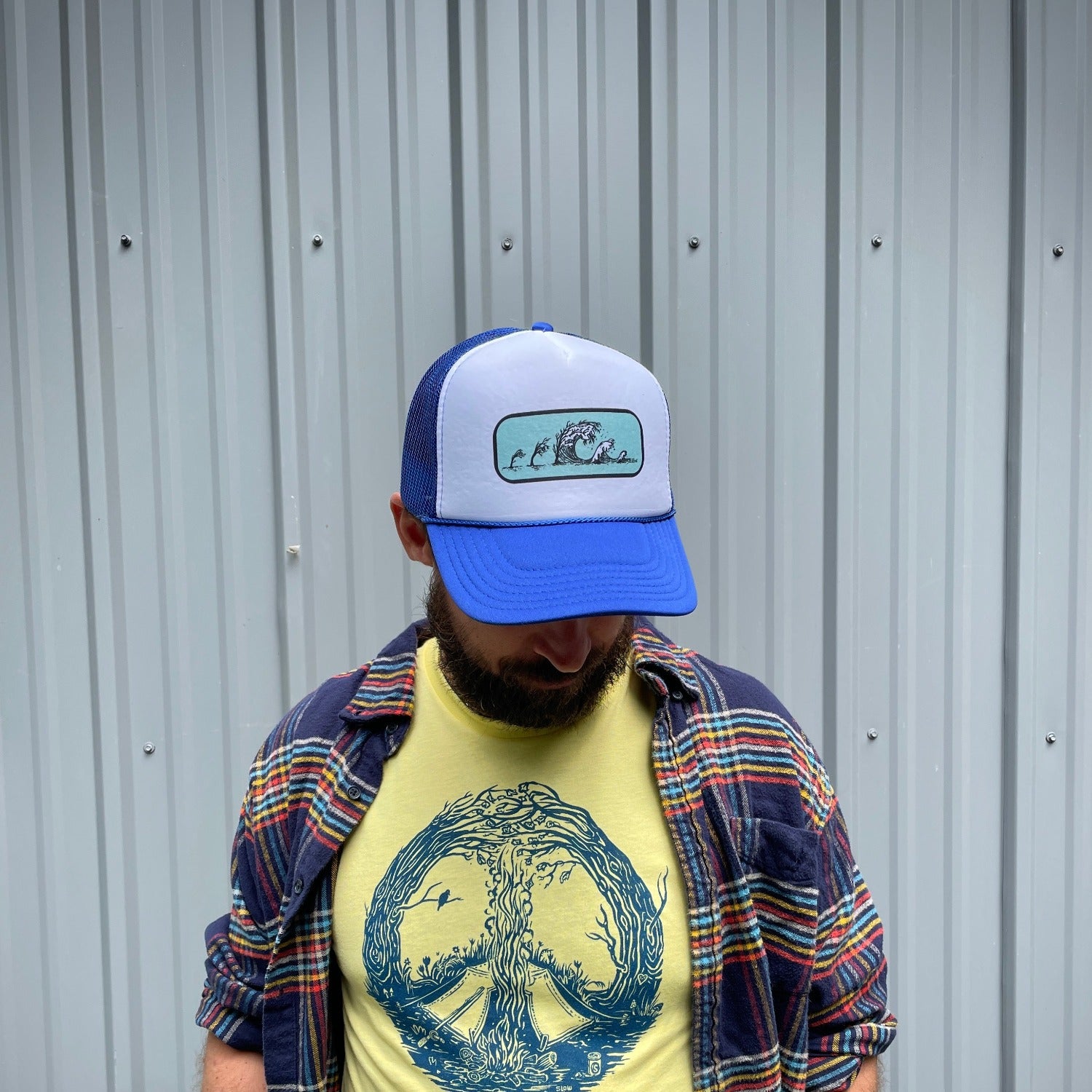 Man wearing t shirt with blue print on a yellow T. Print is of a campfire's smoke going up the center meeting with the tops of curved in trees creating a peace sign. Tent is by the fire and a bird is on a branch