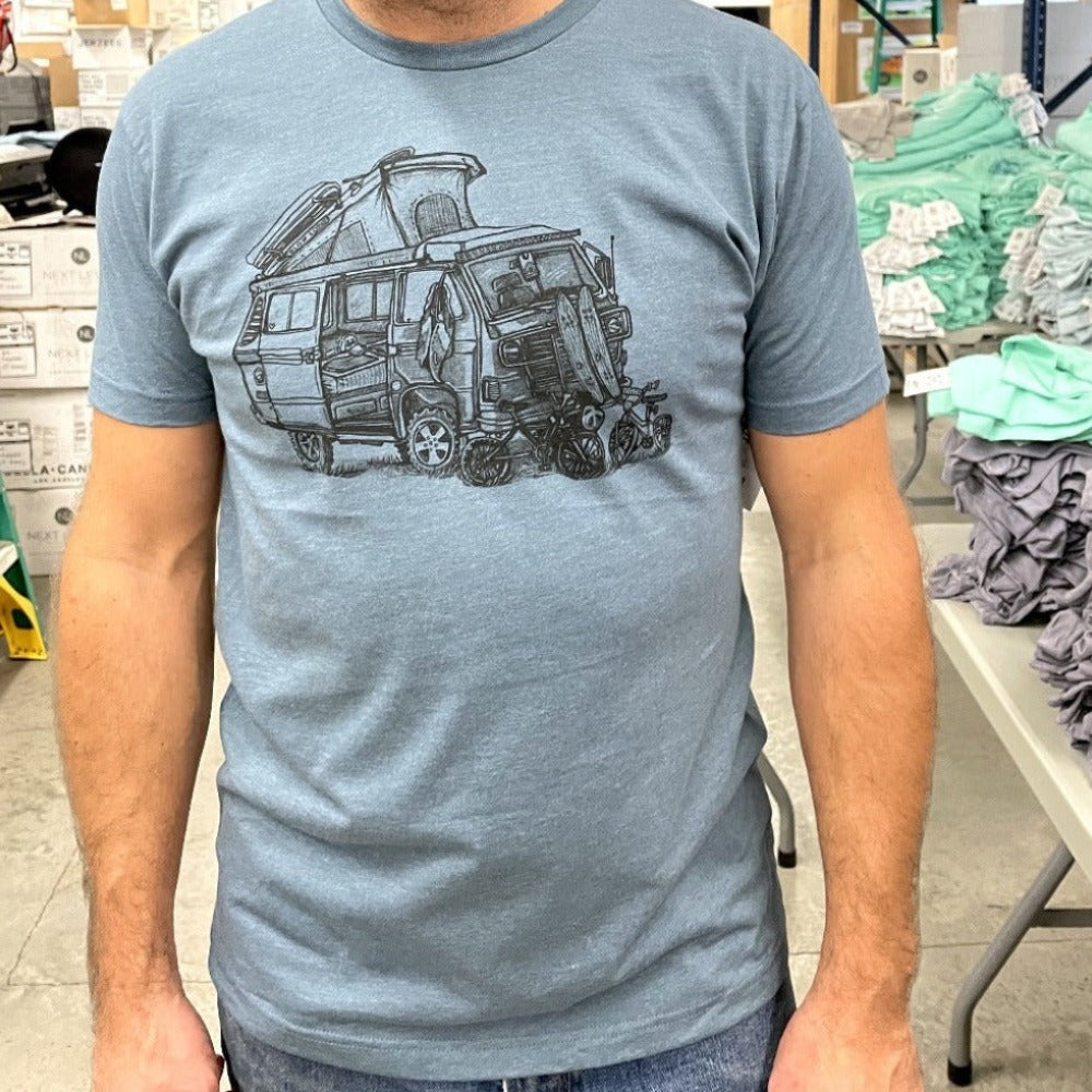 close up of man wearing grey blue shirt with a VW van open and top popped. Skateboards, bikes, and clothing strewn about.