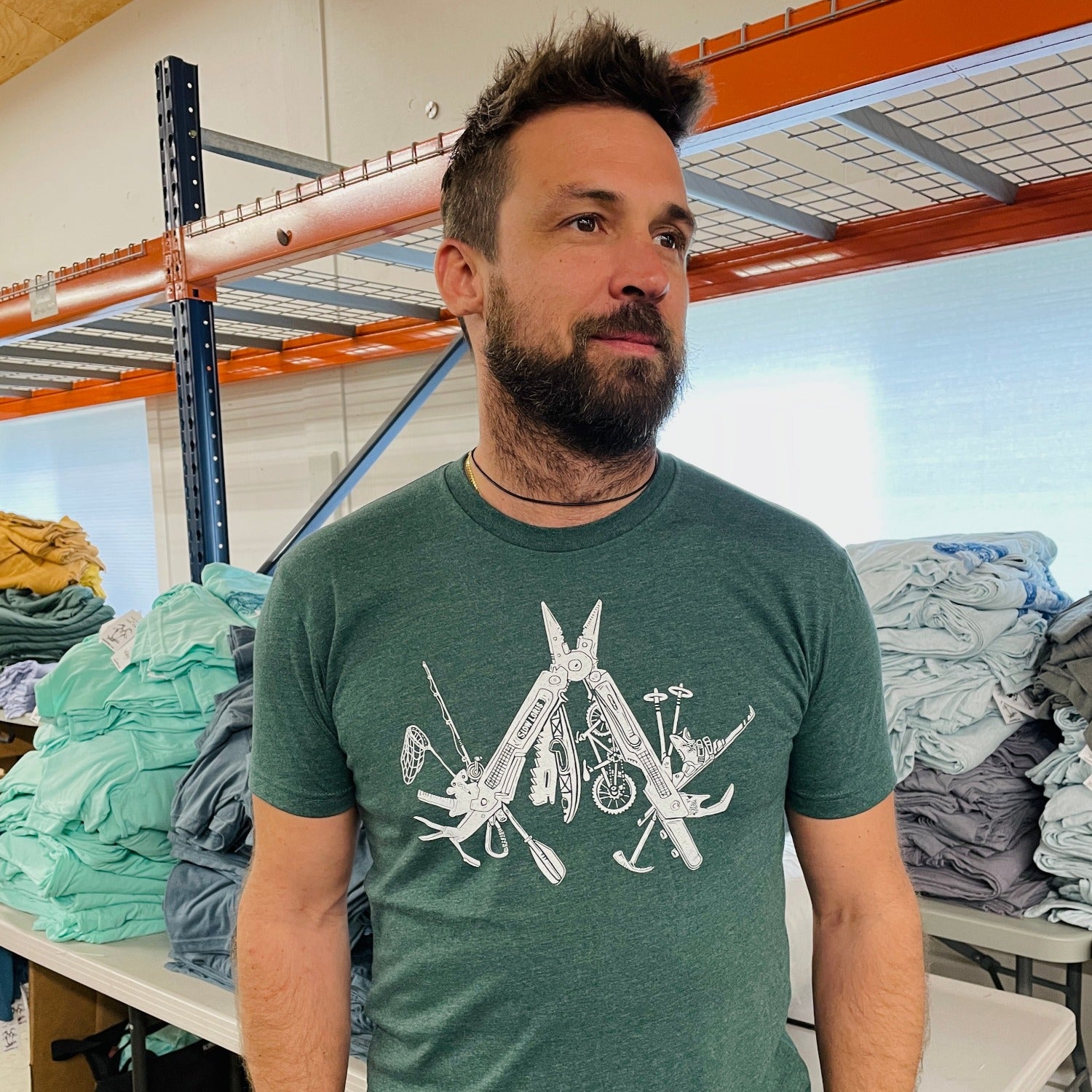man wearing lighter green t-shirt with a white print of a multitool. The tools within the multitool are actually adventuring items- kayak, fishing pole, skis, ice axe, etc etc.